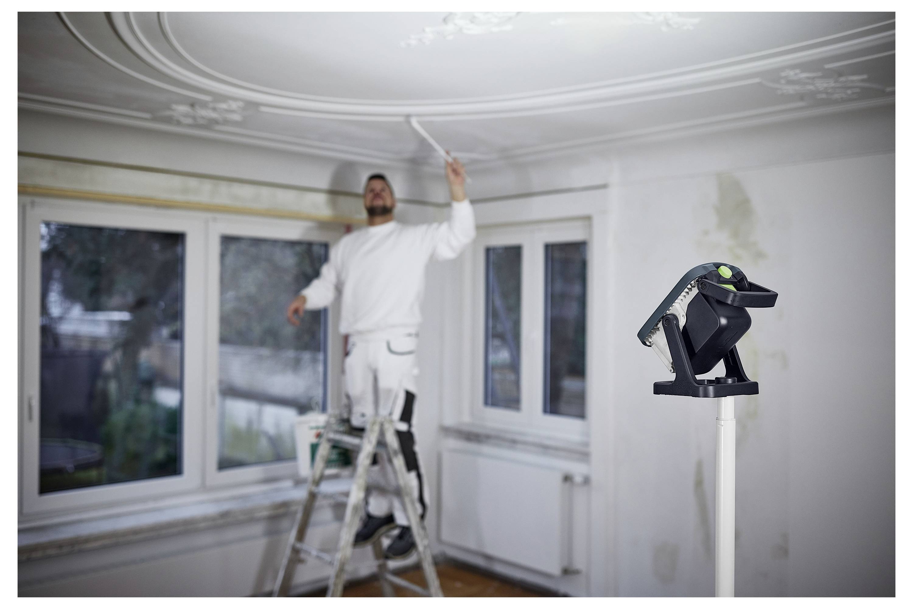 Eine Person steht auf einer Leiter und streicht eine Decke in einem Zimmer mit großen Fenstern. Ein Laser-Nivelliergerät ist im Vordergrund positioniert.