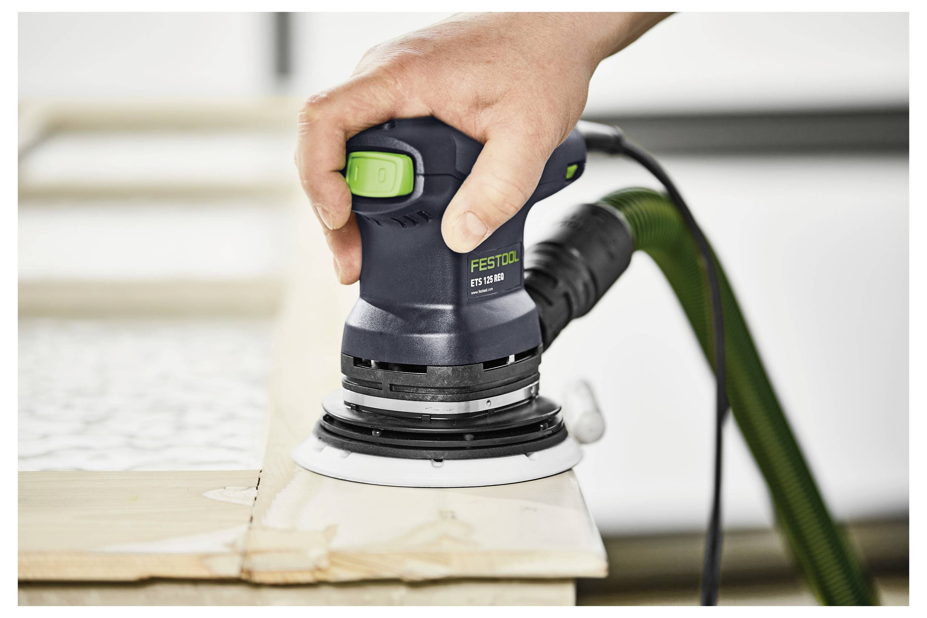 Eine Person verwendet einen Festool-Schleifer, um die Kante einer Holzoberfläche zu glätten, mit einem sichtbaren grünen Schlauch, der zur Staubabsaugung angebracht ist.