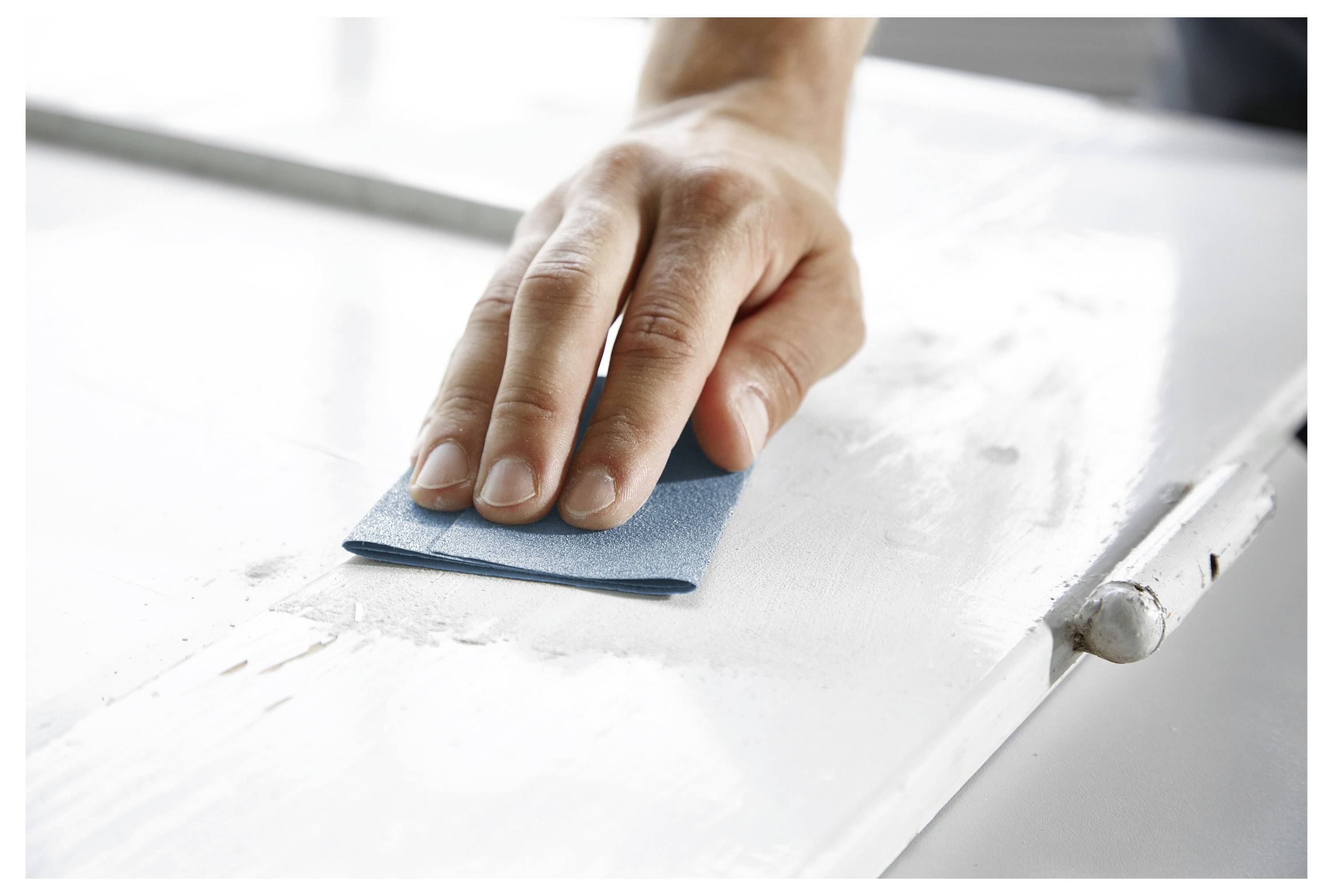 Eine Hand, die Sandpapier verwendet, um eine weiße Holzoberfläche zu glätten, wobei der Fokus auf dem Schleifen liegt.