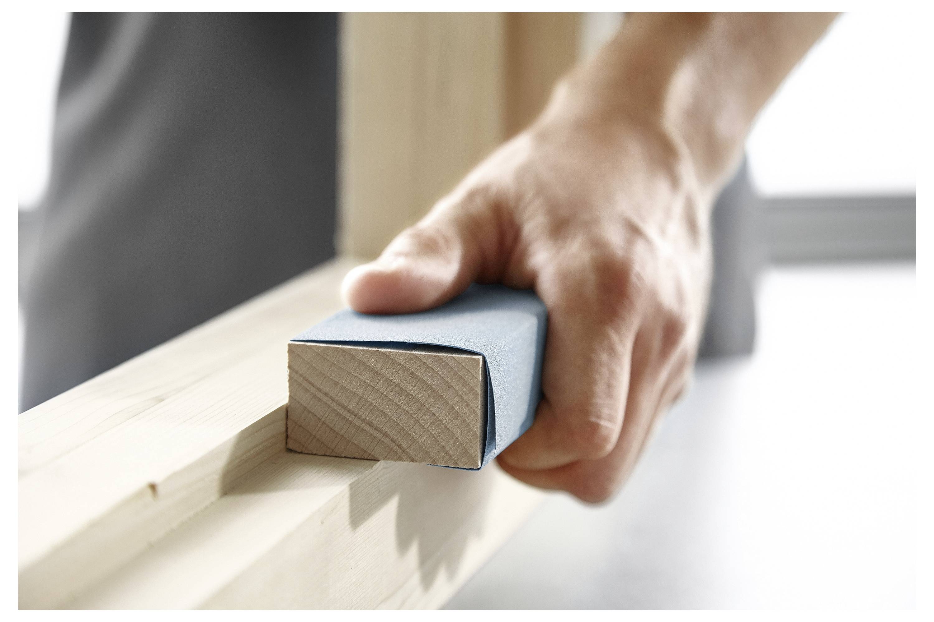 Eine Person, die eine Holzoberfläche mit Schleifpapier abschleifen, wobei der Fokus auf dem Glätten der Kante eines Holzrahmens in einer Werkstattumgebung liegt.
