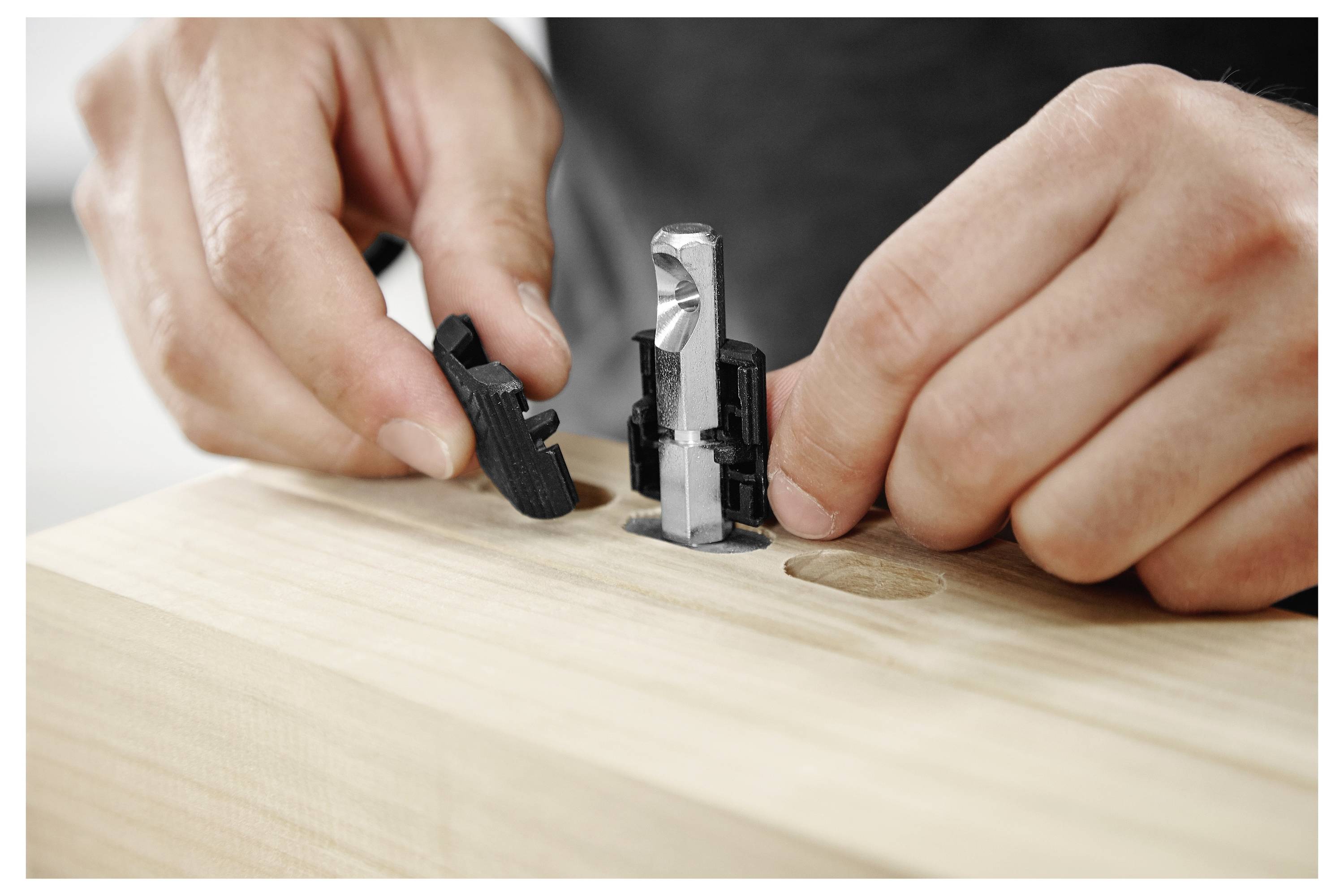 Hände montieren einen schwarz-silbernen Hardware-Stecker in eine Holzoberfläche mit vorgebohrten Löchern, wobei der Fokus auf Konstruktionsdetails liegt.