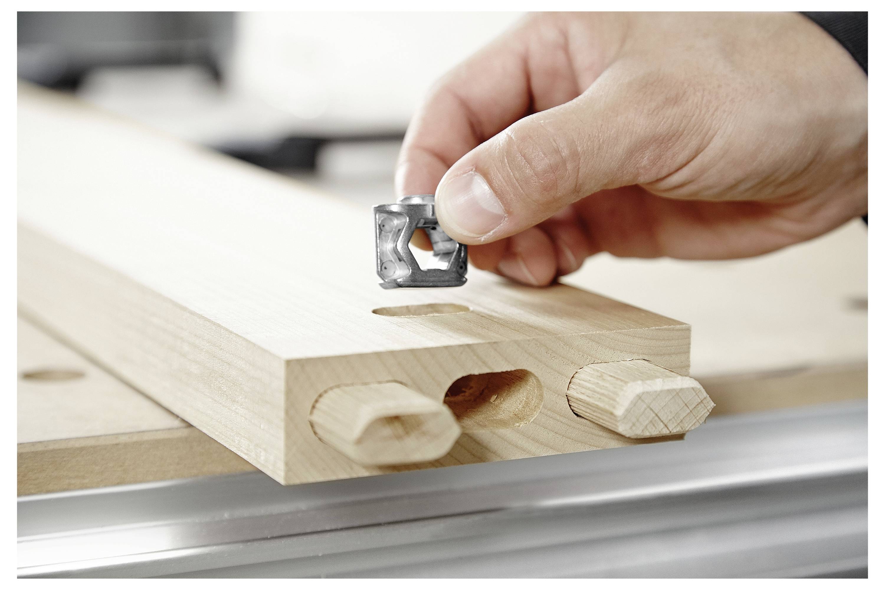 A hand places a metallic tool piece on a wooden board with two dowels inserted, indicating assembly or construction work.
