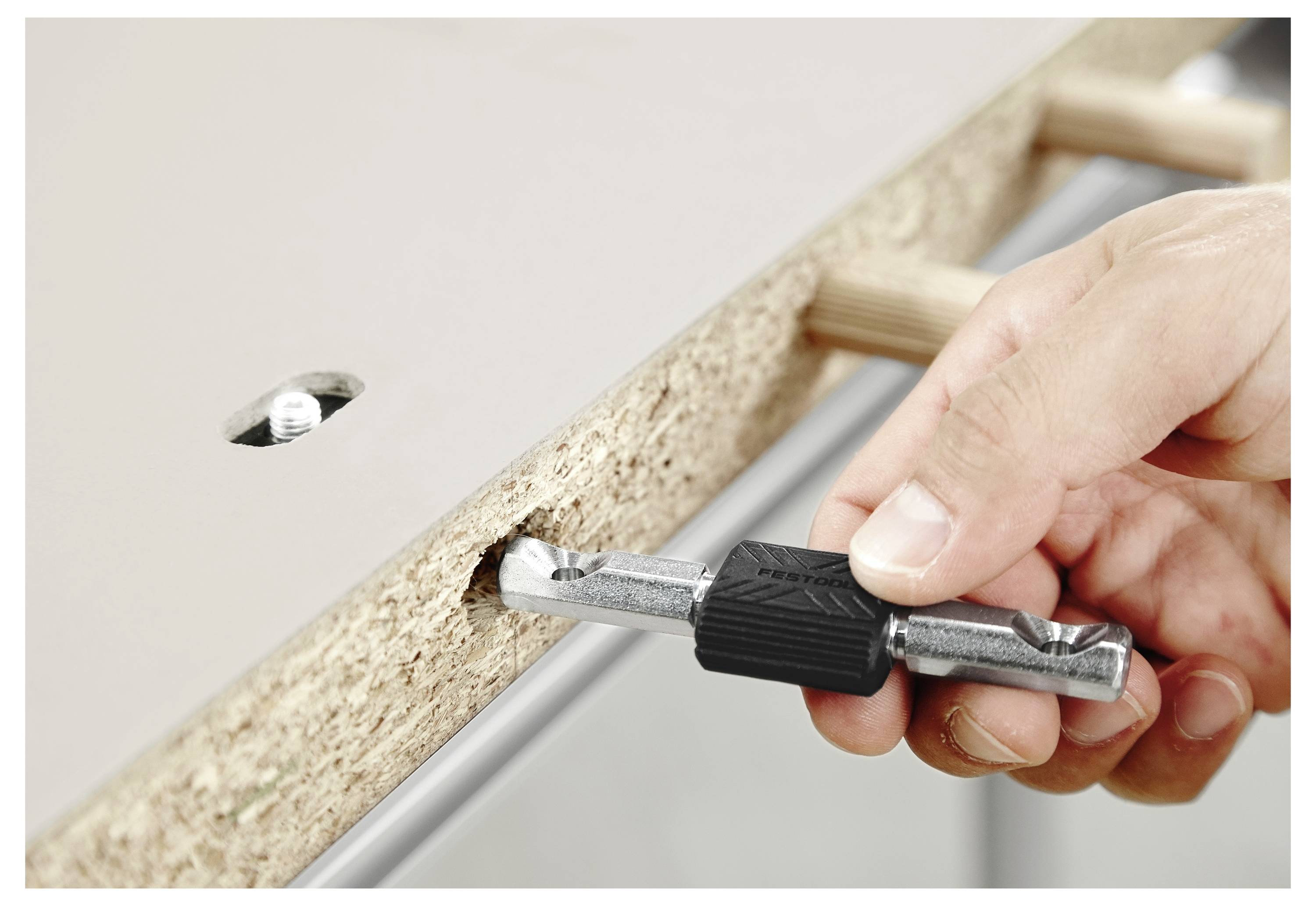 Eine Hand zieht einen Nockenriegel in einem vorgebohrten Loch auf einer Holzplatte fest und montiert Möbelteile mit sichtbaren Holzdübeln und Verbindungselementen.