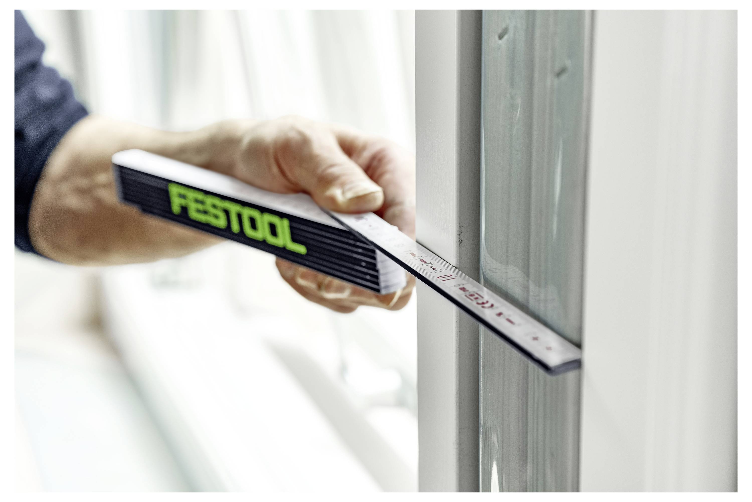 Eine Person verwendet ein Festool-Messinstrument, um die Breite eines Fensterrahmens im Innenbereich zu überprüfen, um eine präzise Passung und Ausrichtung zu gewährleisten.