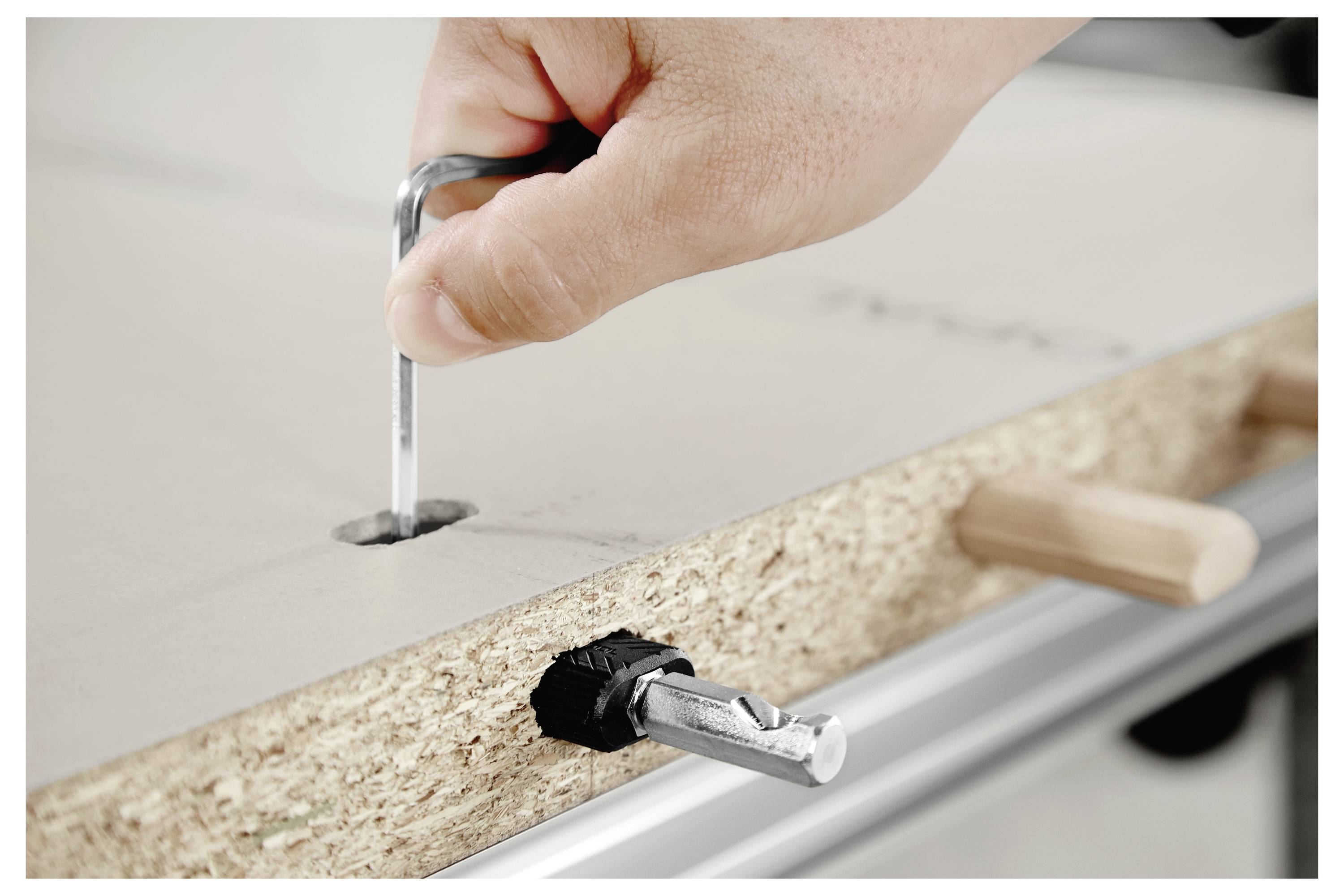 Eine Hand zieht eine Sechskantschraube mit einem Inbusschlüssel in ein Stück Holz, wobei darunter ein Metallverbinder und Holzdübel sichtbar sind.