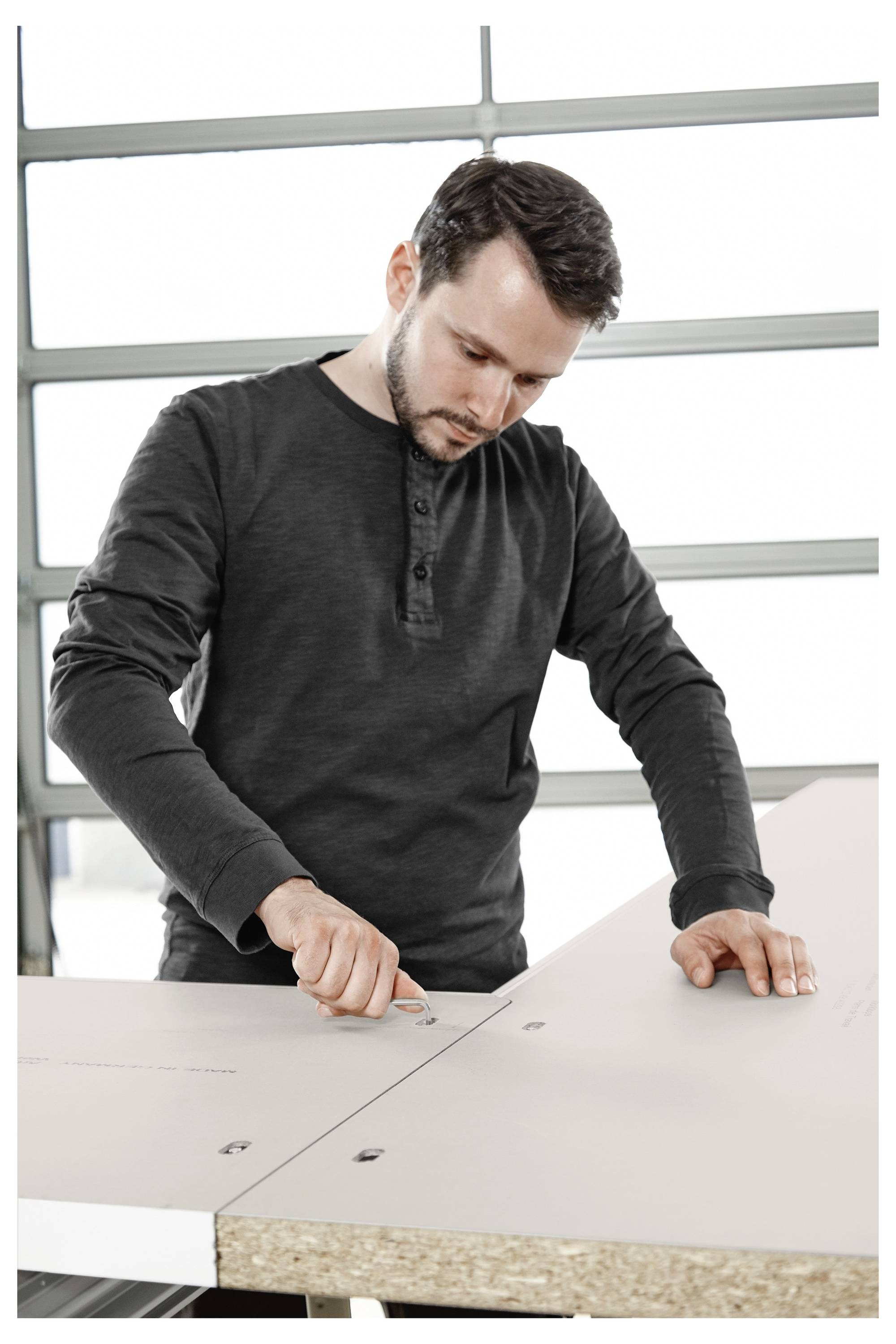 Eine Person, die auf einem großen Brett an einer Werkbank misst und markiert, konzentriert sich intensiv auf die präzise Platzierung. Im Hintergrund eine garagenartige Umgebung.