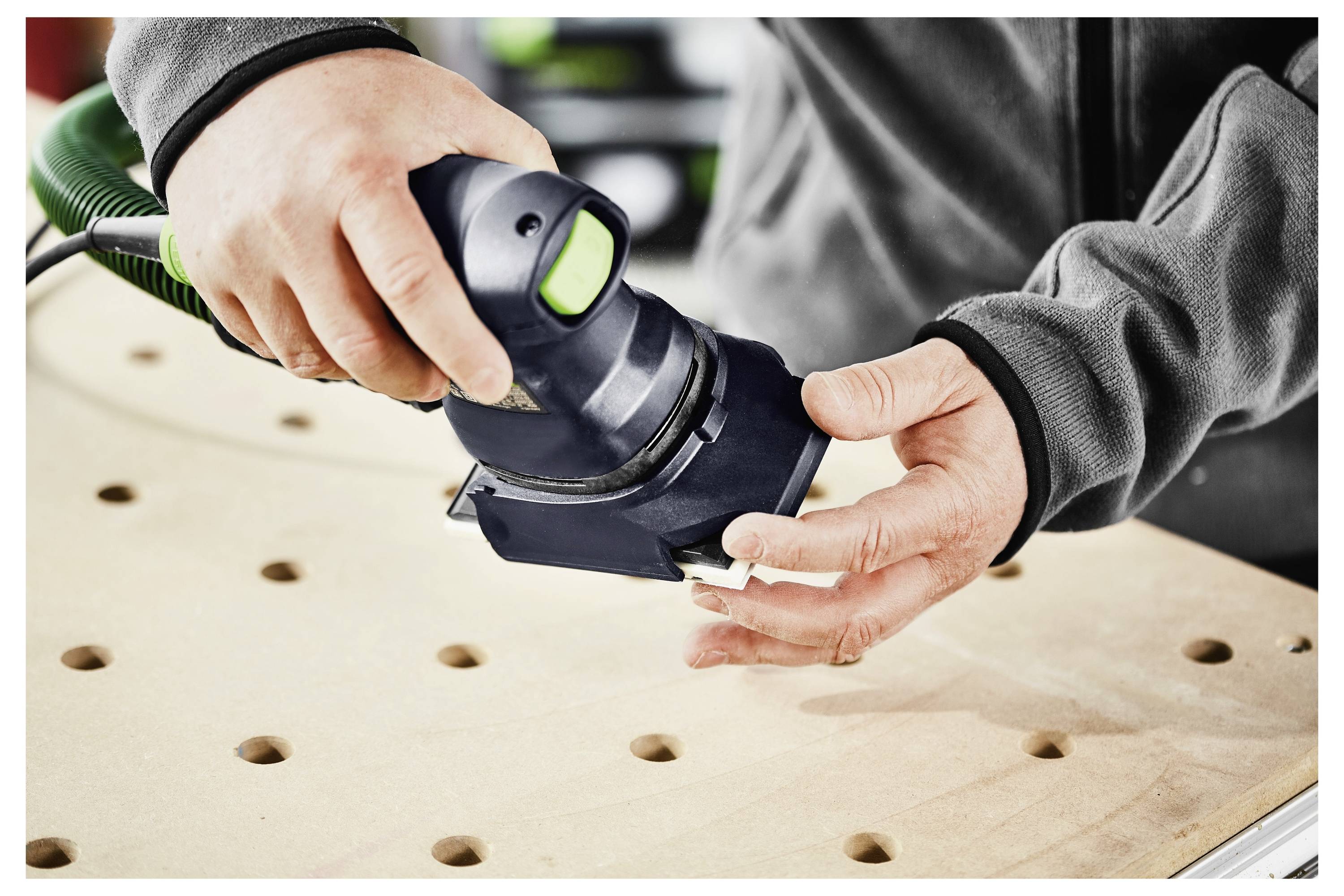 Eine Hand bedient einen Festool-Elektroschleifera auf Holz, mit Fokus auf präzise Handwerkskunst, mit unscharfem Werkstatthintergrund.