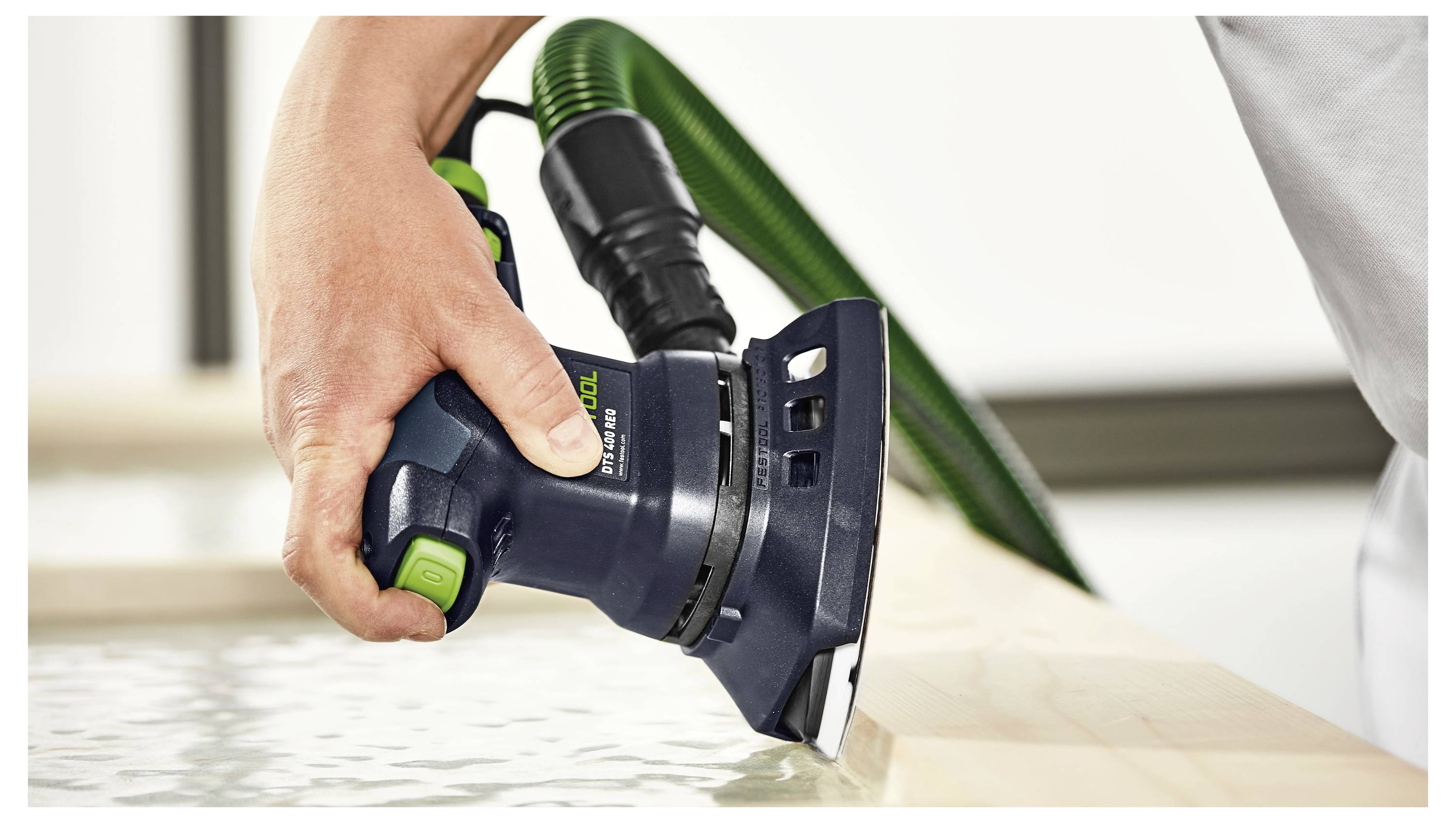 Eine Person verwendet einen elektrischen Schleiffer mit einem grünen Absaugschlauch, um die Kante einer Holzoberfläche zu glätten, wobei der Fokus auf Präzision und Oberflächenfinish liegt.