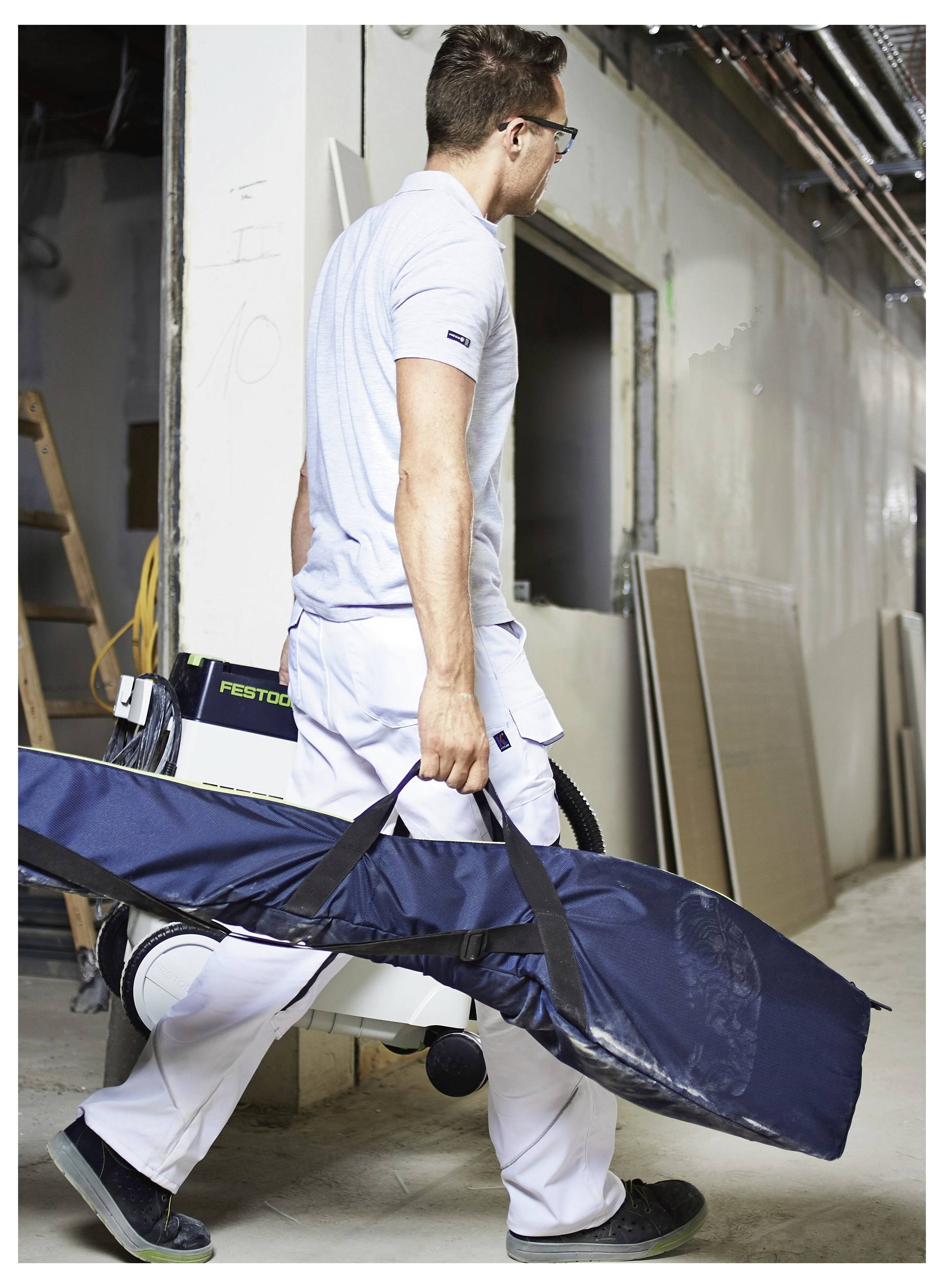 Eine Person in einer Werkstattumgebung trägt einen blauen, länglichen Transportkoffer. Sie trägt weiße Arbeitskleidung und eine Brille und geht an Werkzeugen vorbei.