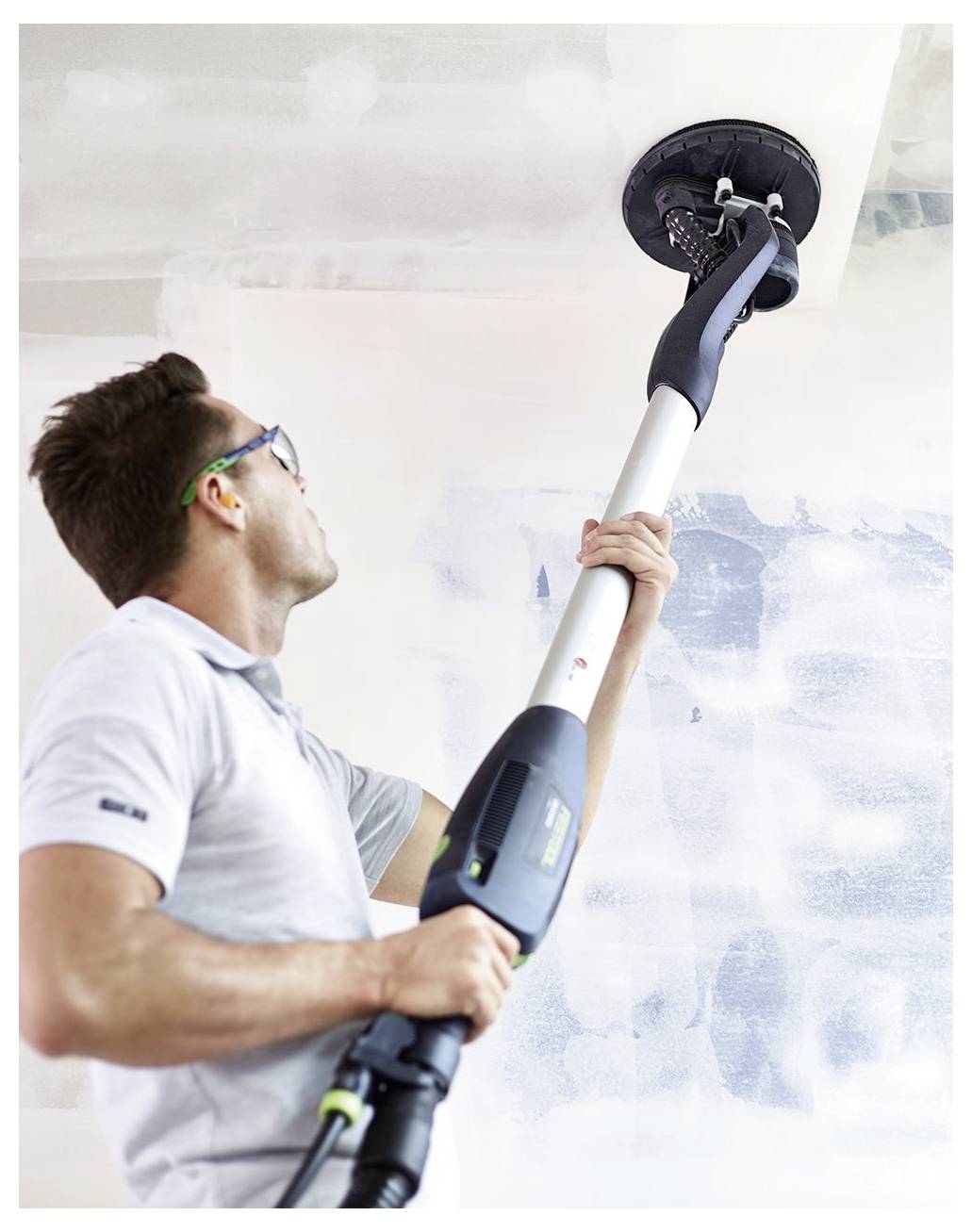 Eine Person in Schutzbrille schleifen eine weiße Decke mit einem langstieligen Elektroschleiffer im Innenraum und konzentriert sich auf das Glätten der Oberfläche.