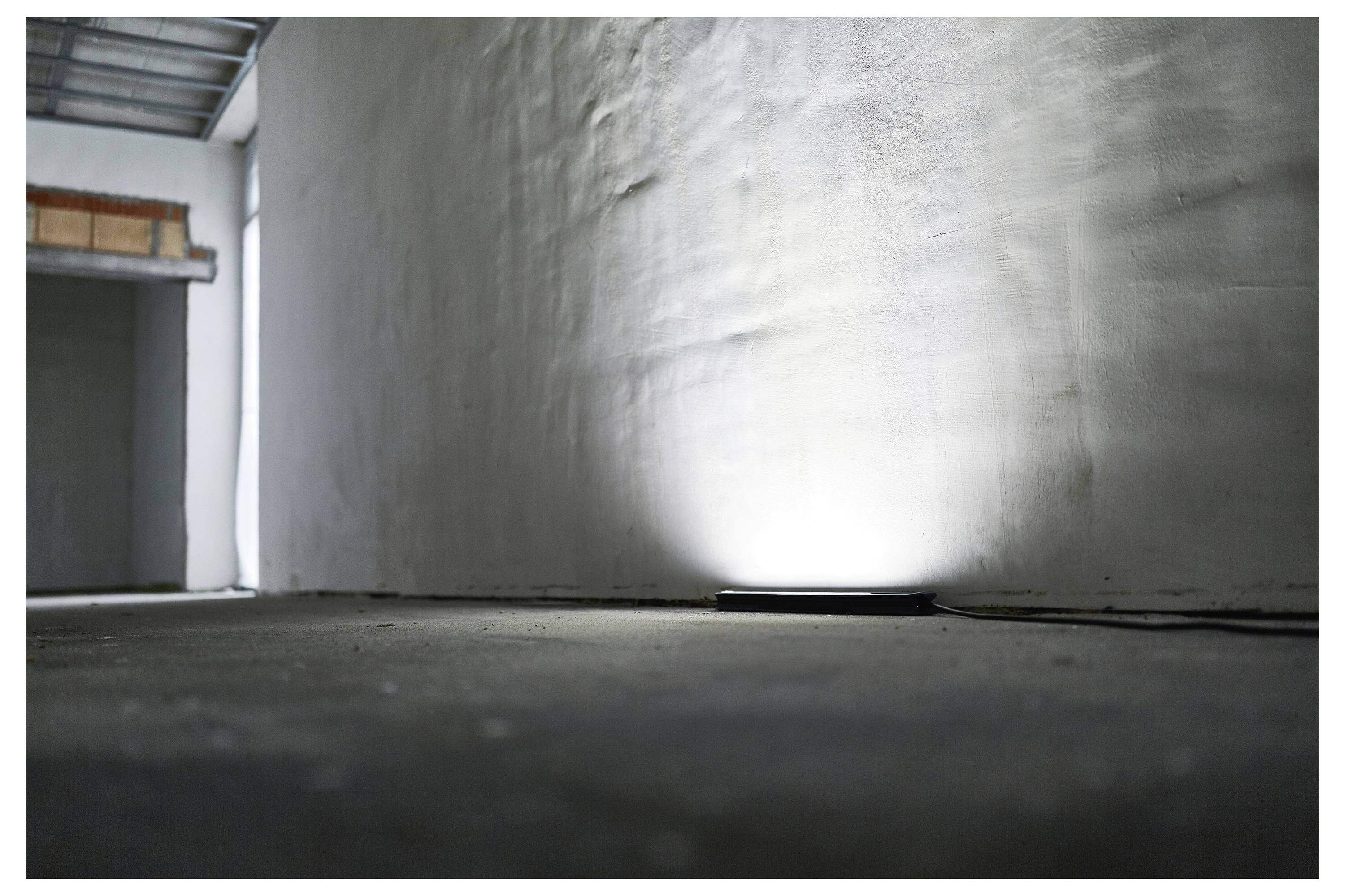 Ein Betonraum mit einem Scheinwerfer auf dem Boden, der Licht auf eine strukturierte Wand wirft und eine minimalistische und nachdenkliche Atmosphäre schafft.