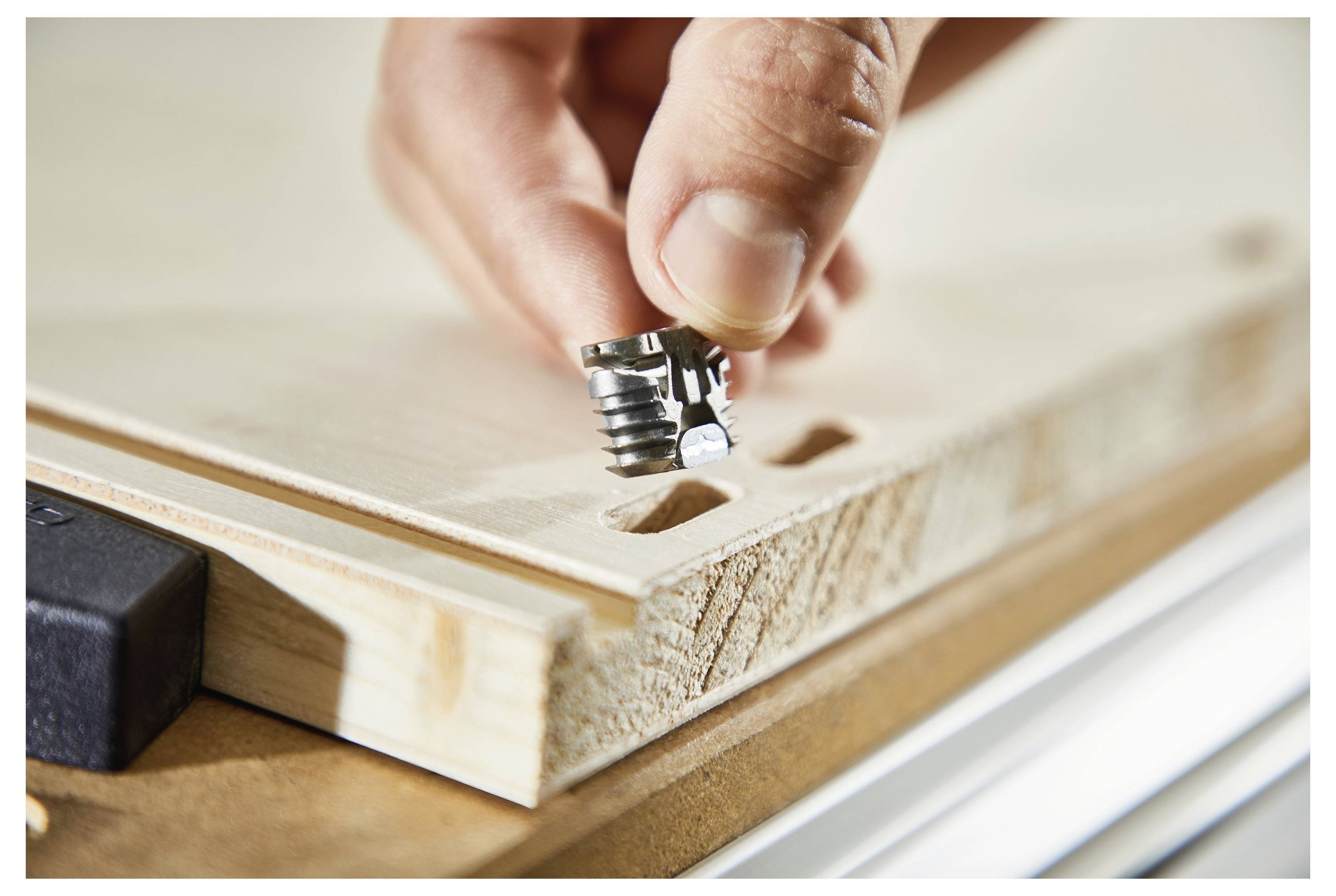 Eine Hand führt einen Metallverbinder in ein Holzbrett mit vorgebohrten Löchern ein, was auf Möbelmontage oder eine Holzbearbeitungstätigkeit hindeutet.