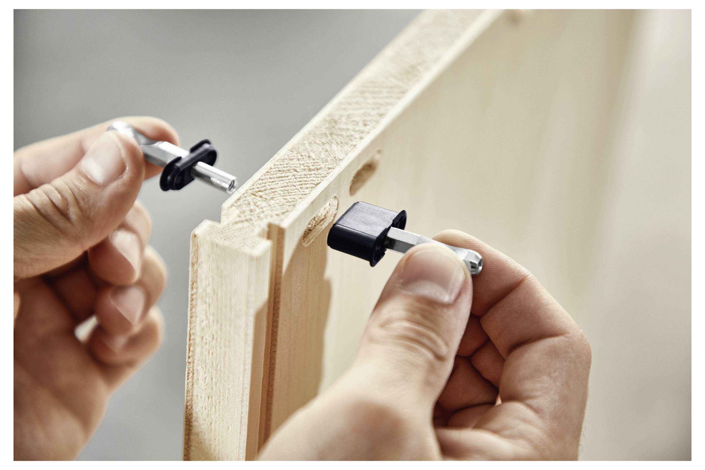 Hände montieren ein Holzstück mit Exzenterverschlüssen und richten diese in zwei gebohrte Löcher aus, um Möbelteile zu befestigen.