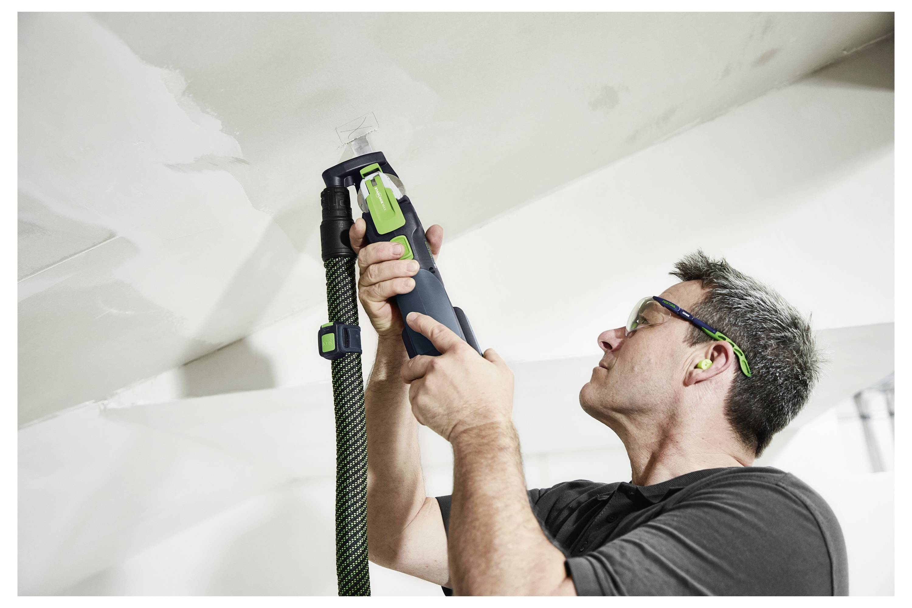 Ein Mann mit einer Schutzbrille benutzt ein Elektrowerkzeug mit einem Staubschlauch, um eine Decke zu schleifen, und konzentriert sich intensiv auf die Aufgabe.
