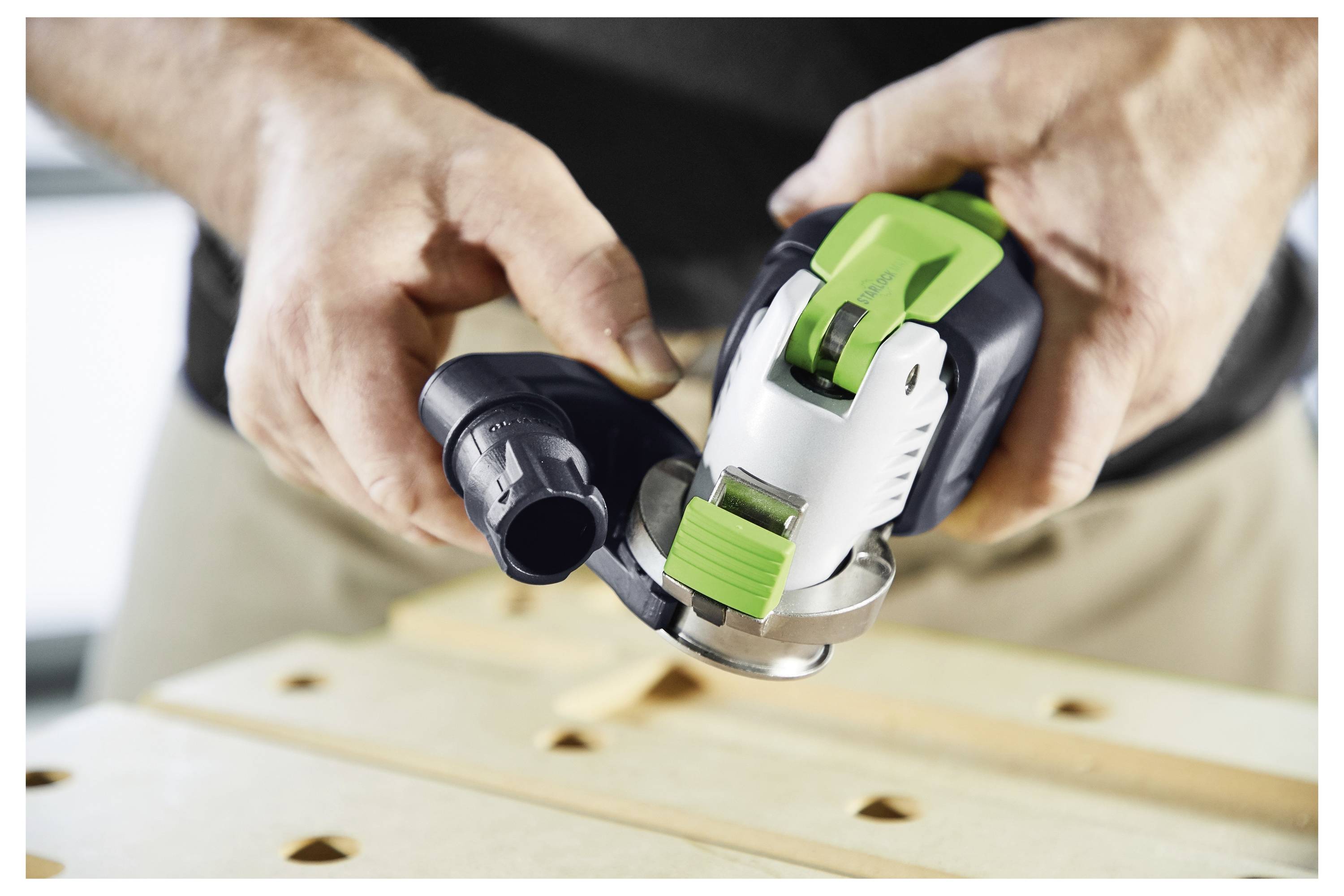 A person is holding a cordless green and black handheld tool, possibly a sander, over a wooden surface in a workshop setting.