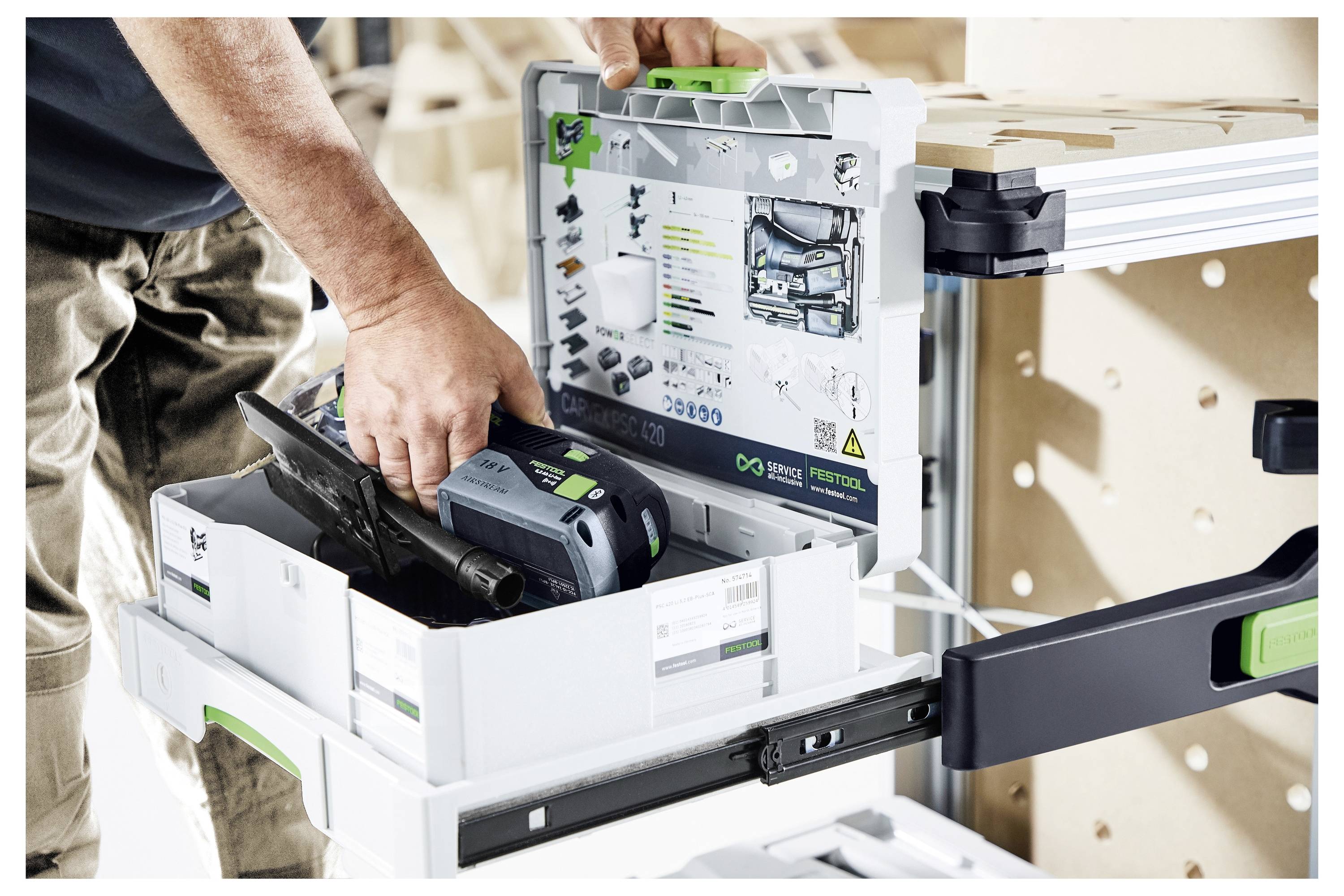 Eine Person organisiert ein Elektrowerkzeug in einer beschrifteten Aufbewahrungsbox in einer Werkstatt. Die Box steht auf einem ausziehbaren Regal, das an einer Werkbank befestigt ist.