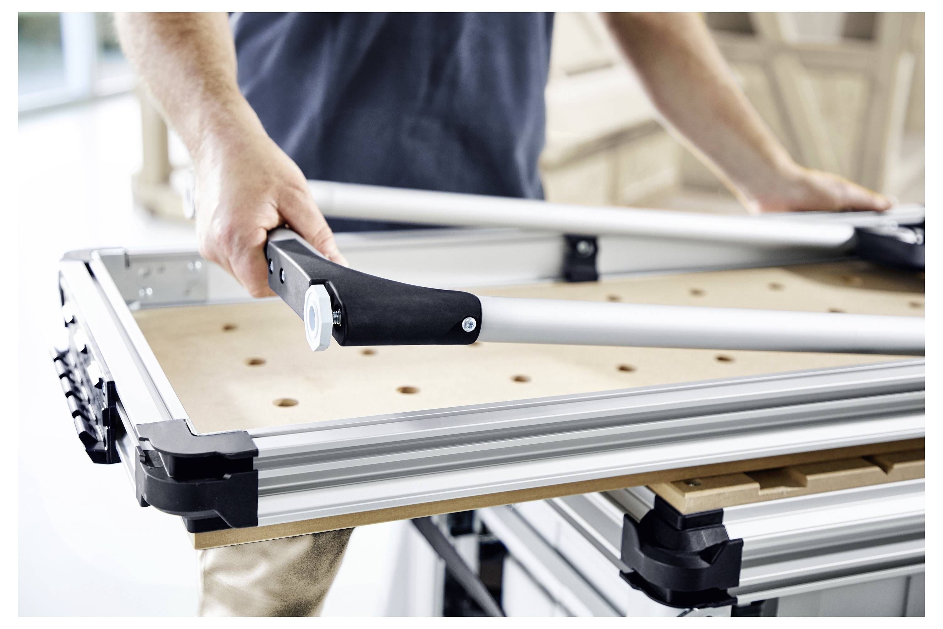 Eine Person, die eine Werkbank mit Aluminiumprofilen und einer Holzoberfläche zusammenbaut, hält ein Metallwerkzeug in einem hellen Arbeitsbereich.