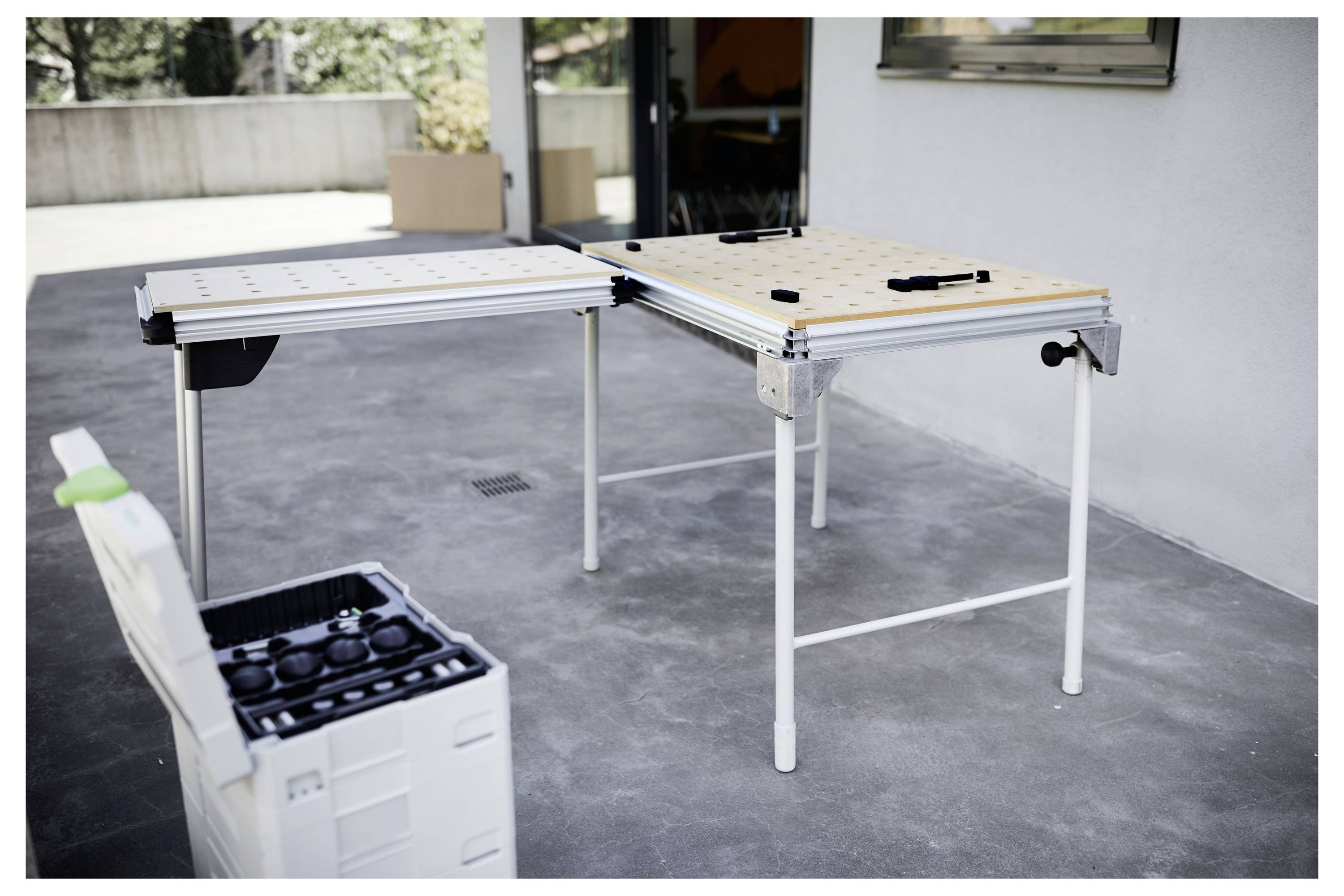 Zwei modulare Werkbänke mit Werkzeugen auf einer Terrasse, neben einer Werkzeugkiste mit verschiedenen Beschlägen. Außenbereich mit Pflanzen im Hintergrund.