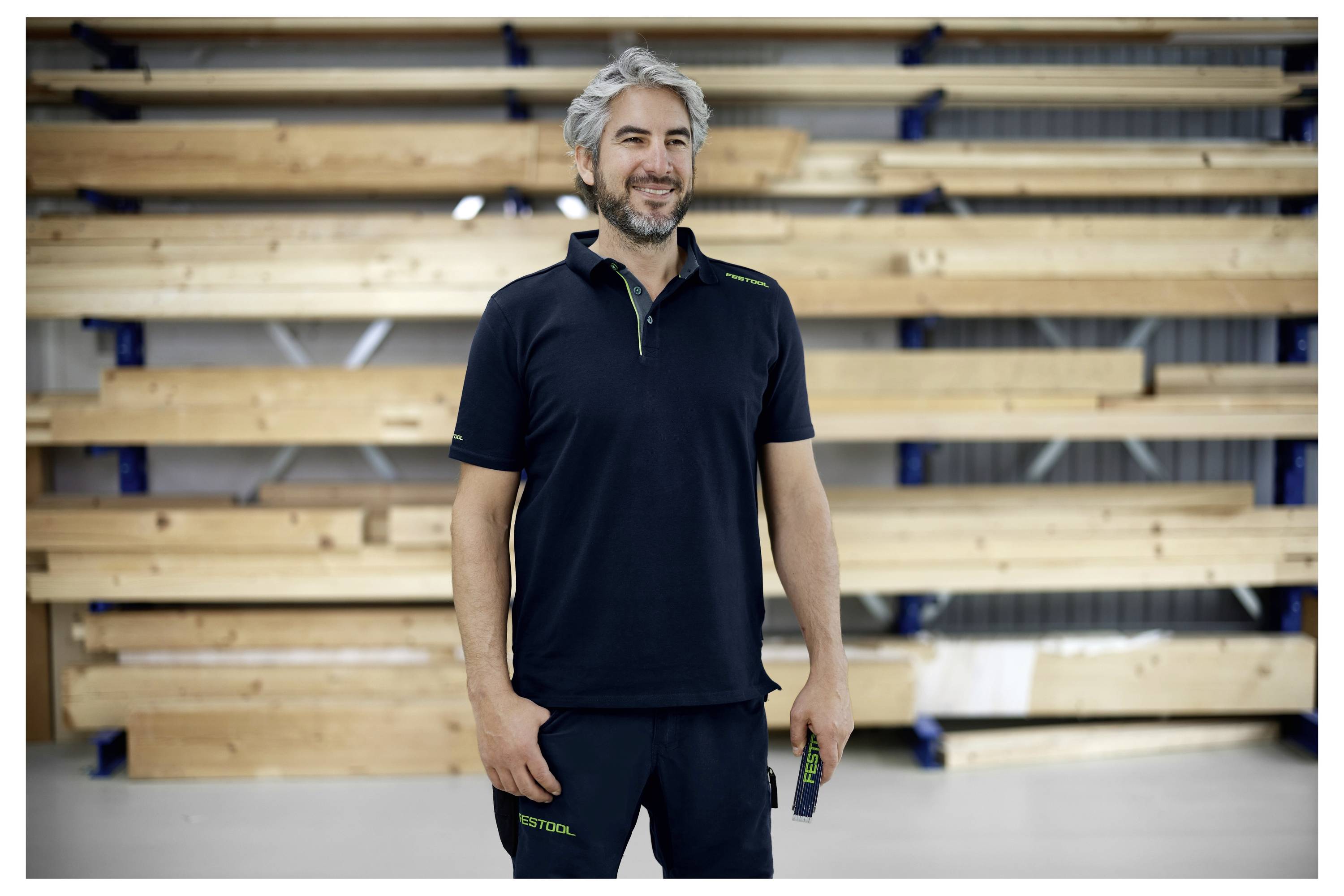 Ein lächelnder Mann in einem schwarzen Poloshirt steht vor gestapelten Holzbrettern, was auf ein Tischler- oder Baugewerbe hindeutet.