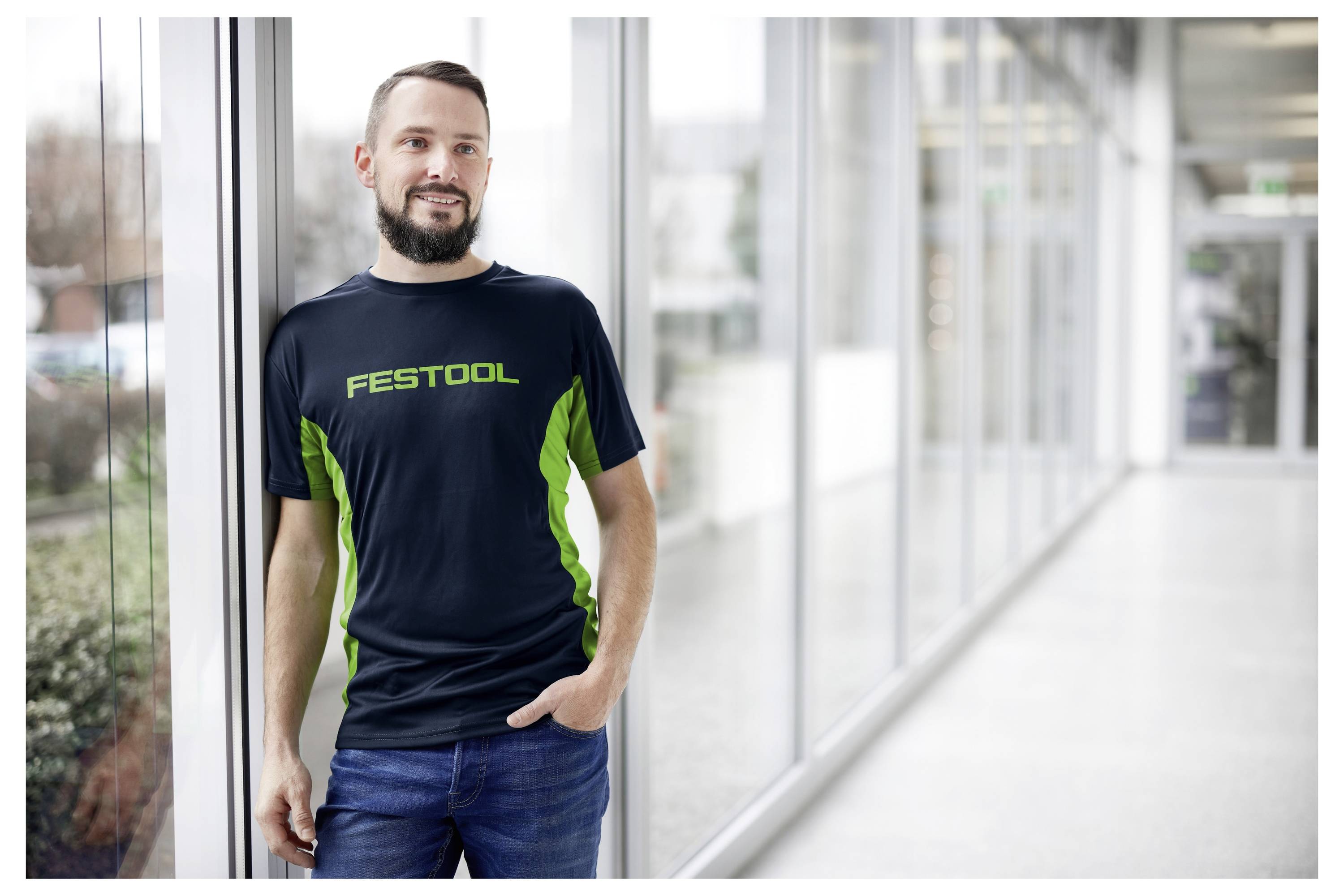 Eine Person steht in einem Flur und lehnt sich gegen eine Wand. Sie trägt ein schwarzes und grünes T-Shirt mit dem Wort 'Festool' auf der Vorderseite.