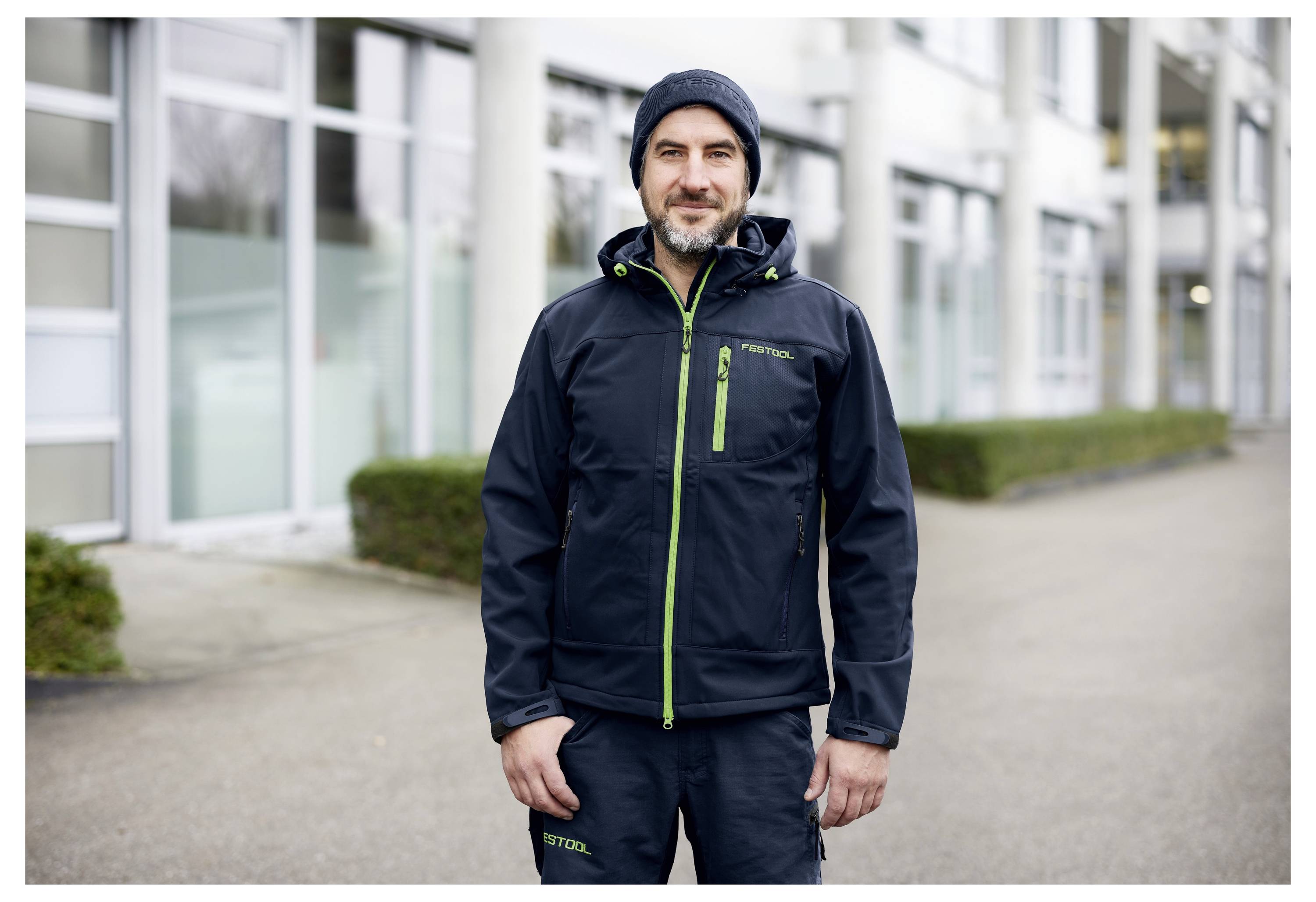 Ein Mann in Outdoor-Sportkleidung steht lächelnd vor einem modernen Gebäude mit großen Fenstern und Säulen. Er trägt eine Beanie und eine Jacke.