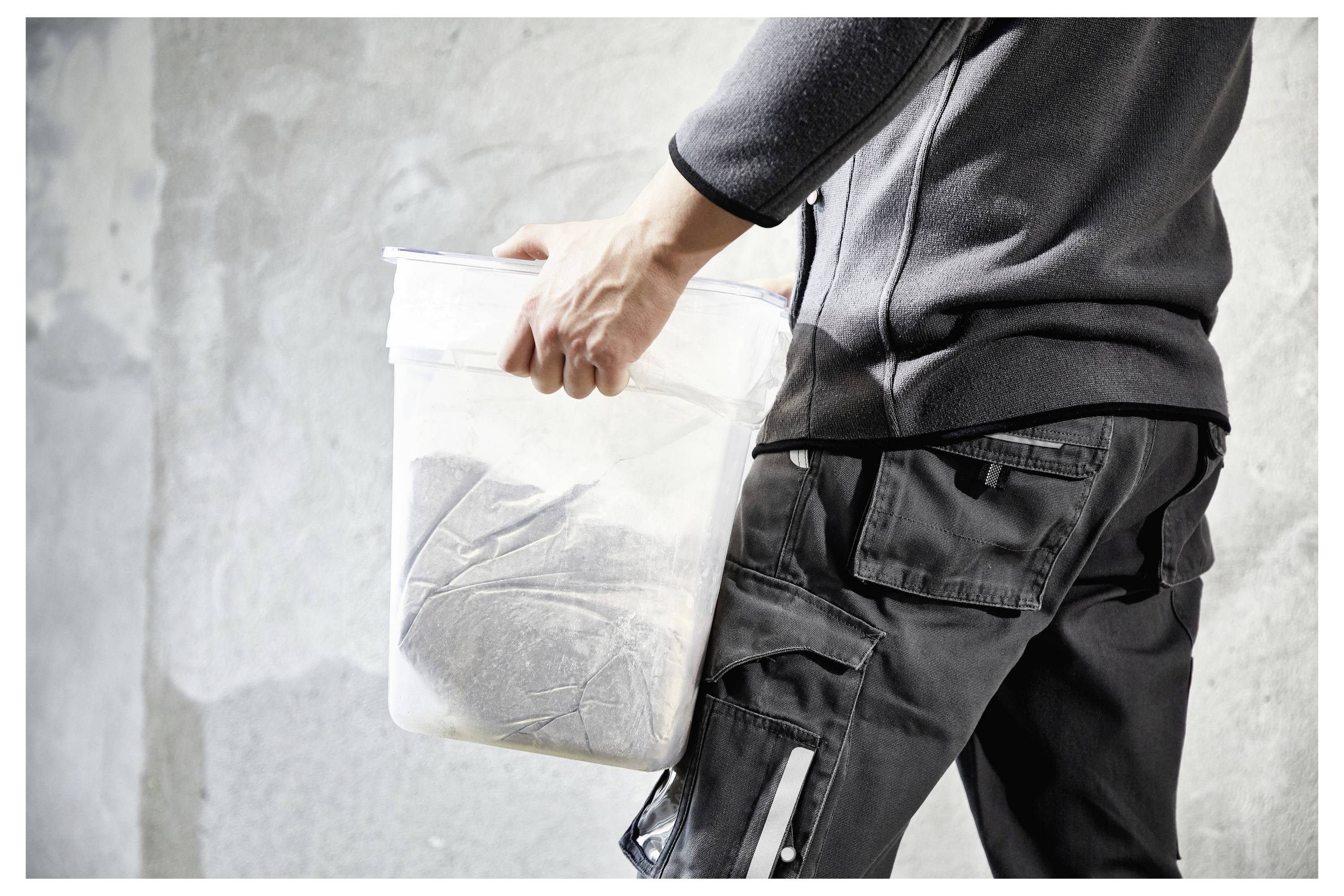Eine Person in einem grauen Outfit trägt einen halbdurchsichtigen Behälter gefüllt mit Zementmischung vor einem strukturierten Wandhintergrund.