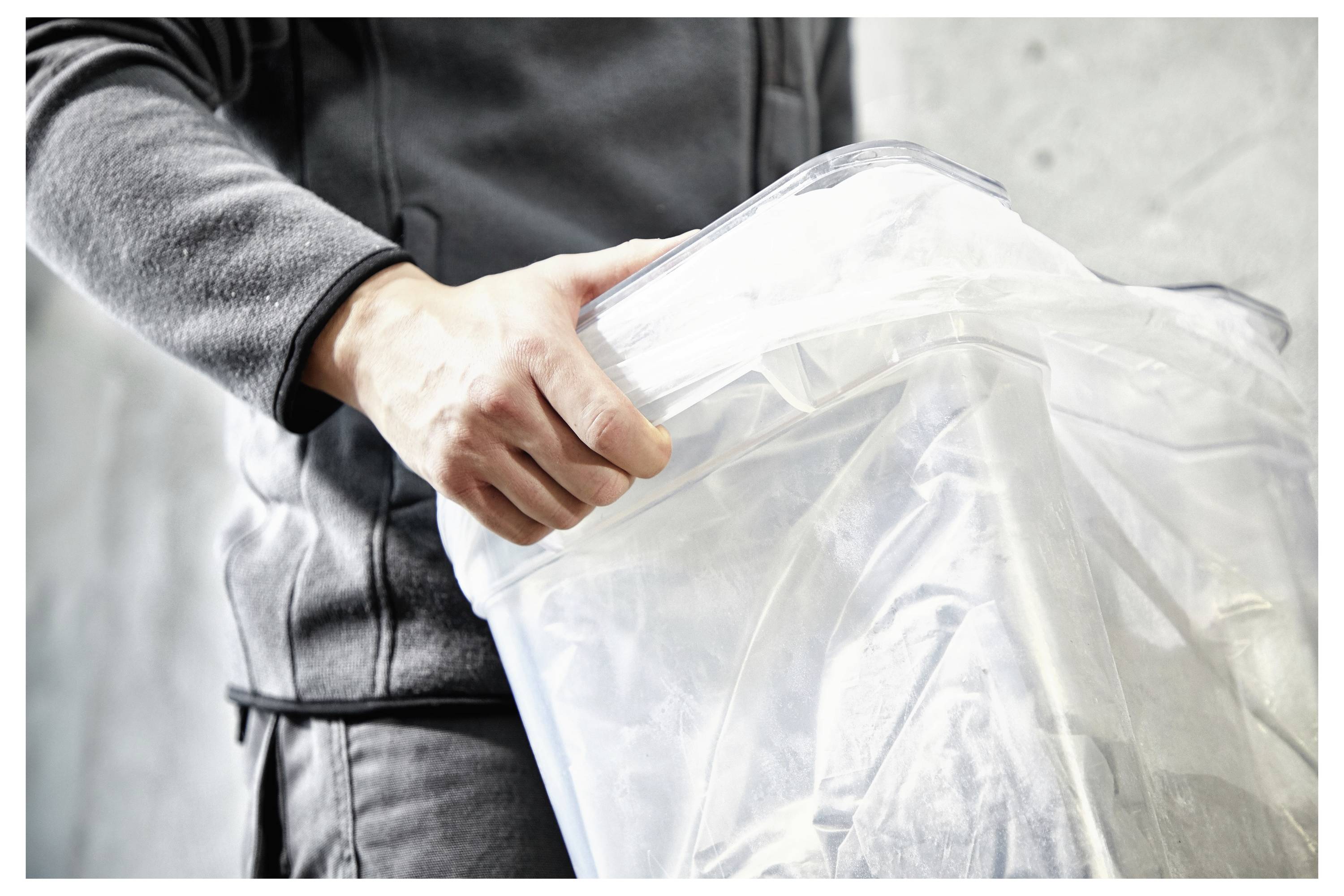 Eine Person trägt eine dunkle Jacke und hält einen durchsichtigen Kunststoffbehälter mit weißem Deckel, was auf Ordnung oder Lagerung in einer häuslichen oder bürolichen Umgebung hindeutet.