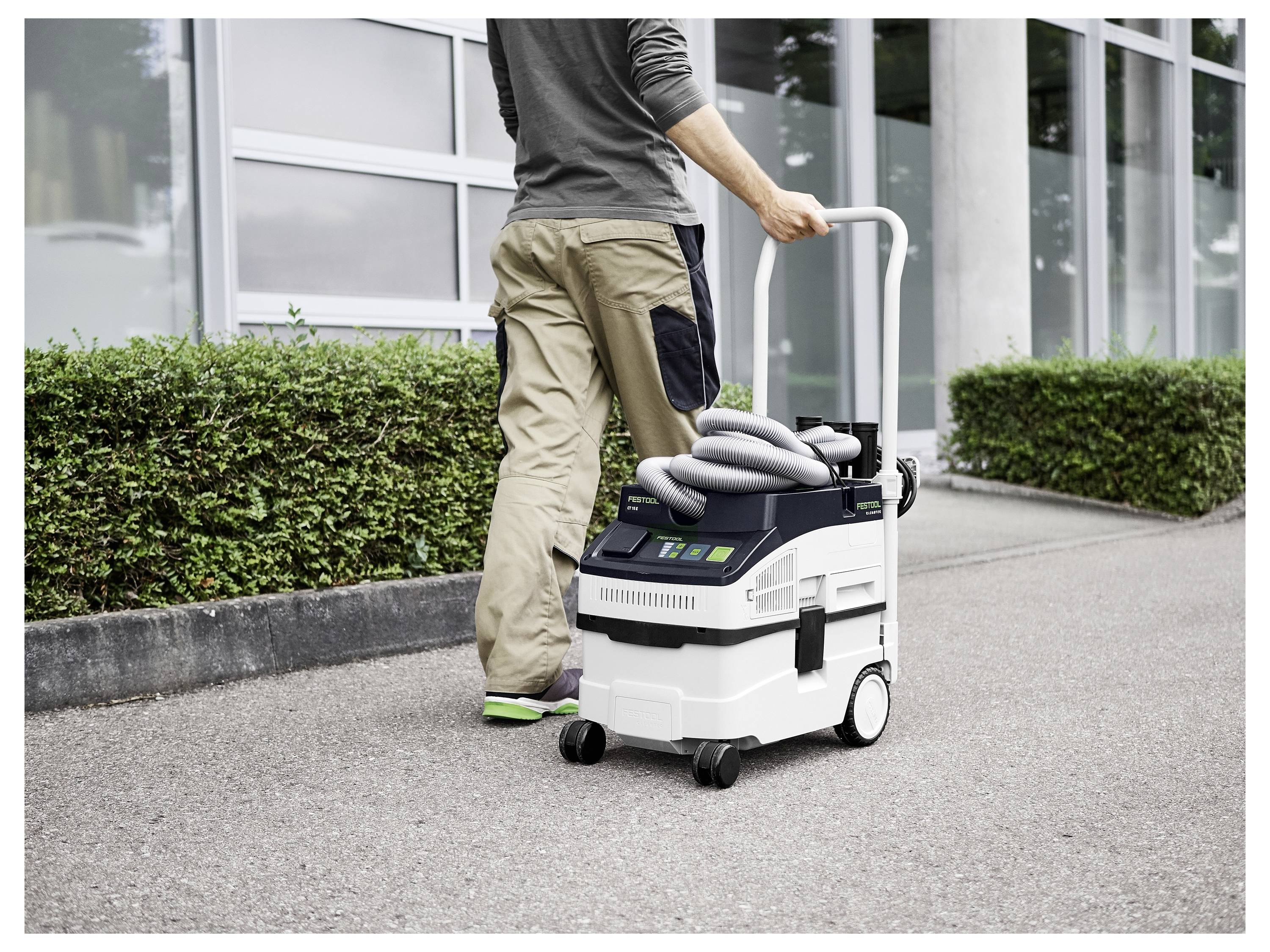 Eine Person schiebt einen tragbaren Staubsauger mit aufgerolltem Schlauch auf einem Gehweg neben einem Gebäude und zeigt die Nutzung im Freien und die Mobilität.