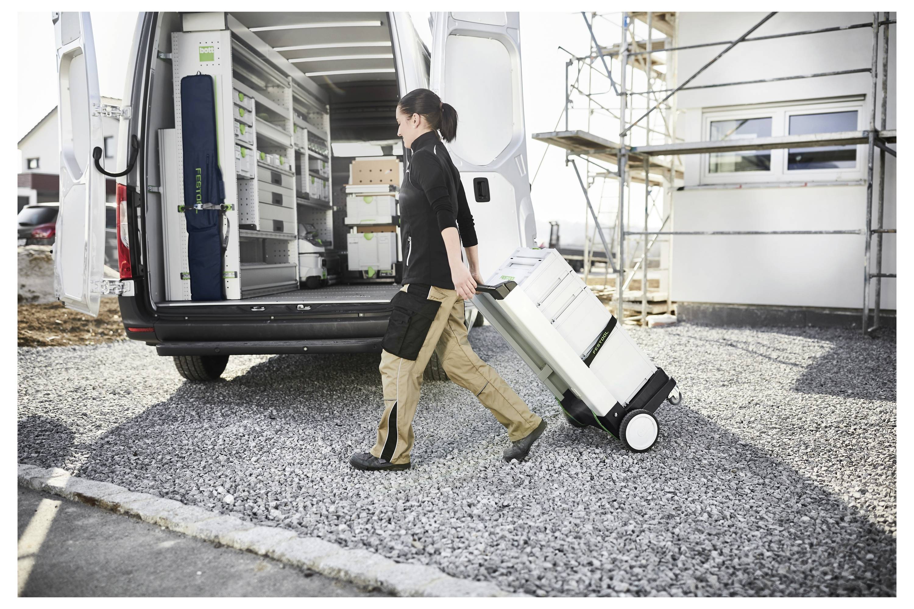 Eine Person zieht eine Werkzeugkiste mit Rädern aus einem Lieferwagen, der auf einer Baustelle in einem Wohngebiet geparkt ist, was Organisation und Vorbereitung in der Arbeitsumgebung andeutet.