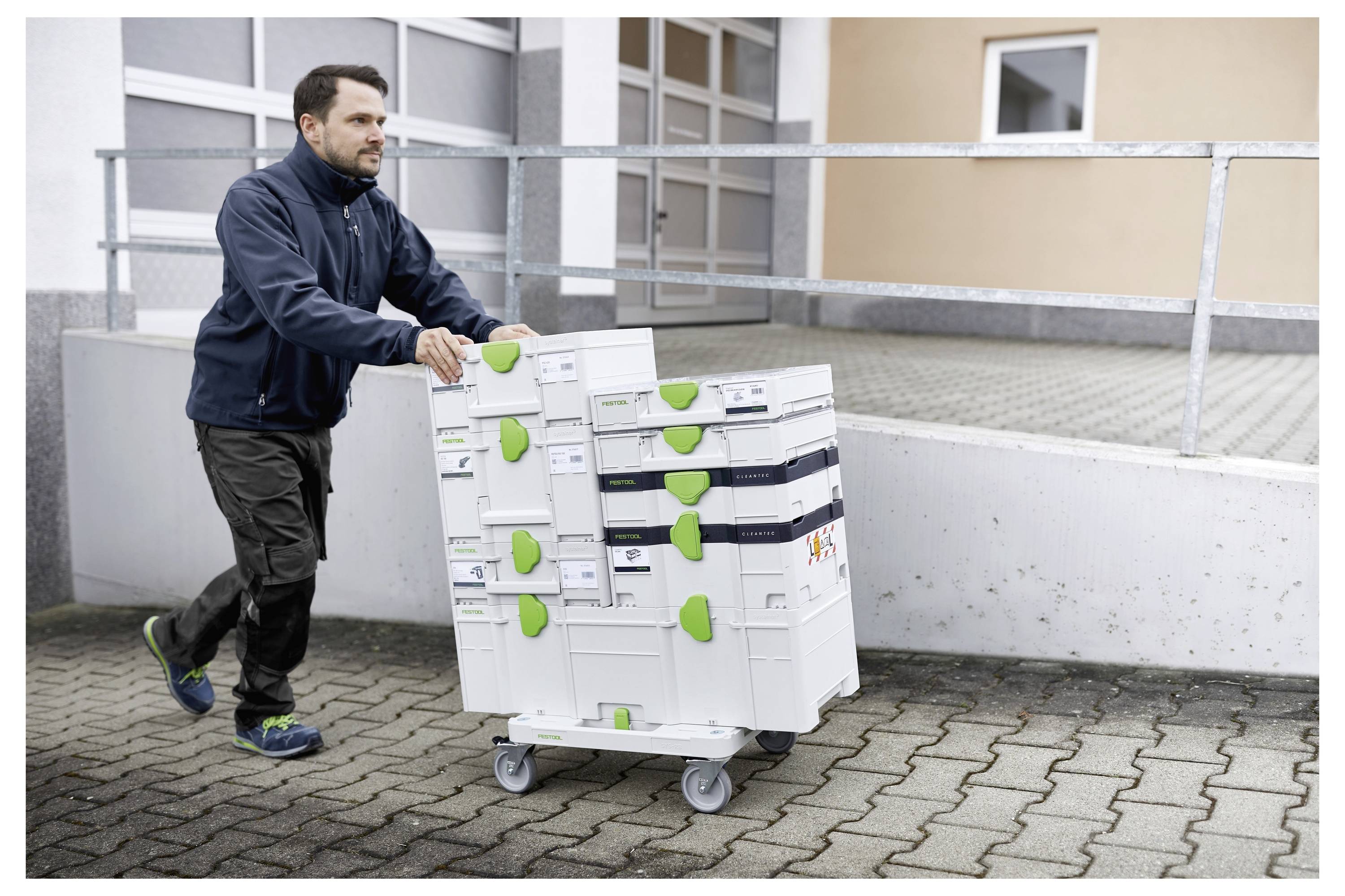 Ein Mann schiebt einen Stapel von Ausrüstungskoffern auf einem Wagen mit Rädern über ein gepflastertes Gebiet in der Nähe eines Gebäudes, was auf einen Transport oder eine Lieferung hindeutet.