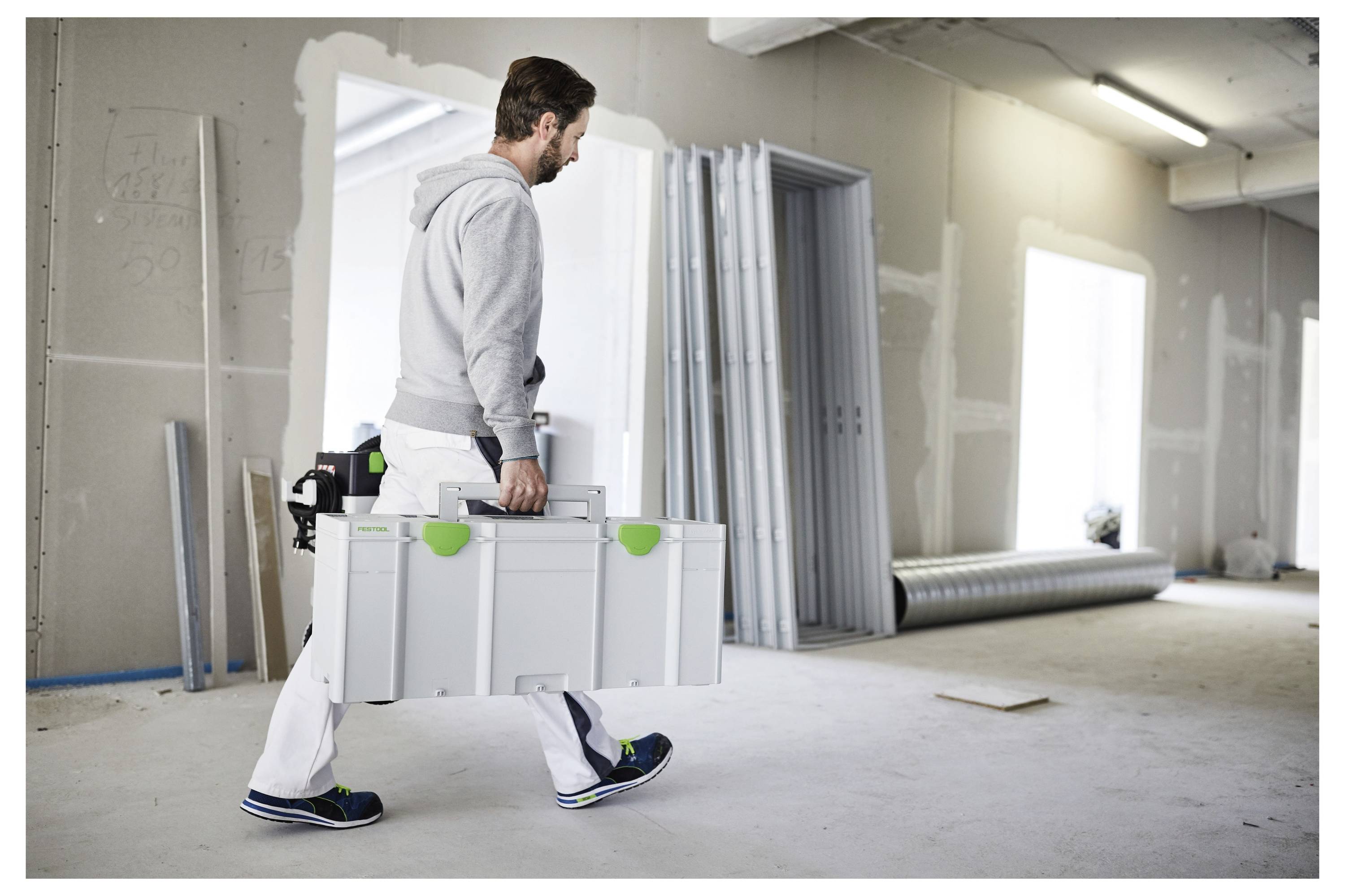 Eine Person in Arbeitskleidung trägt eine große, weiße Werkzeugkiste in einem Raum, der sich im Bau befindet. Die Wände sind unfertig und Baumaterialien sind sichtbar.