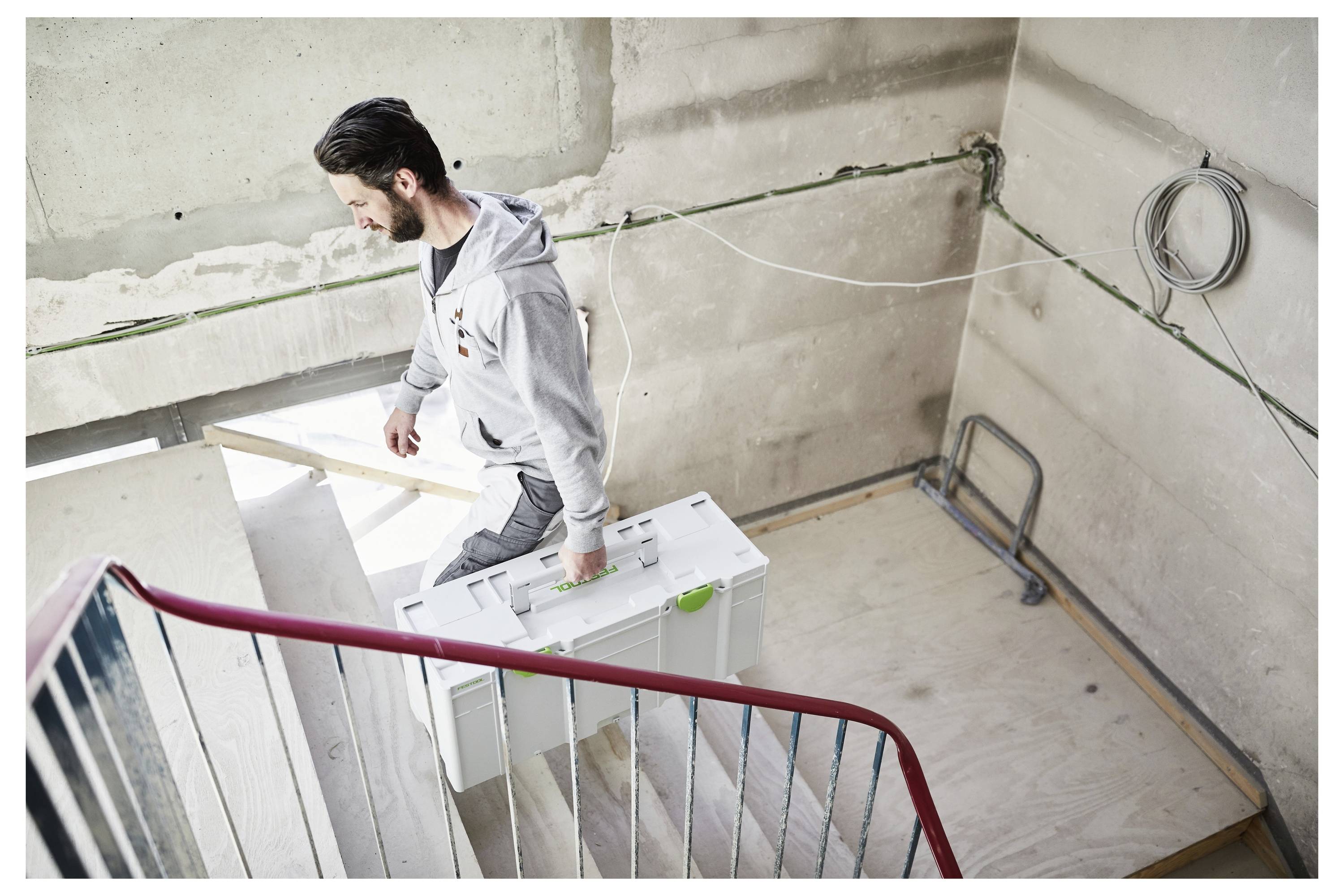 Ein Mann trägt eine große weiße Schachtel eine Betontreppe in einem unfertigen Gebäude hinauf, was auf Bau- oder Renovierungsarbeiten hindeutet.