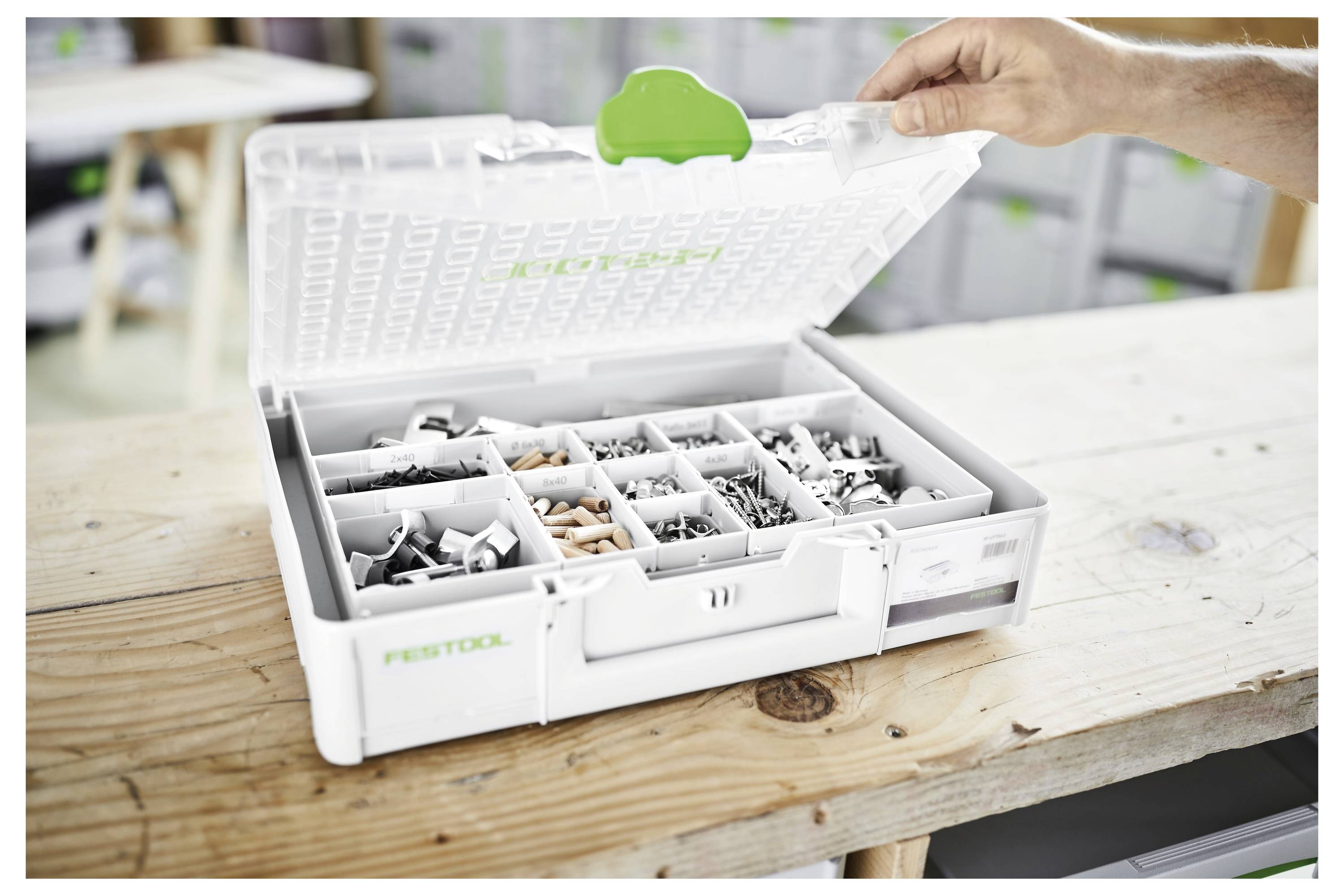 Eine offene Werkzeug-Organizer-Box mit verschiedenen Schrauben und Kleinteilen steht auf einem Holztisch. Eine Hand hebt den Deckel.