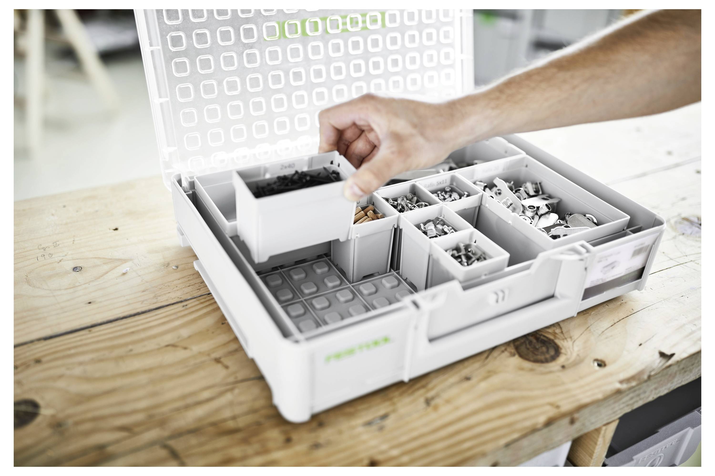 Eine Hand organisiert Schrauben und kleine Metallteile in einer unterteilten Aufbewahrungsbox auf einem Holztisch.