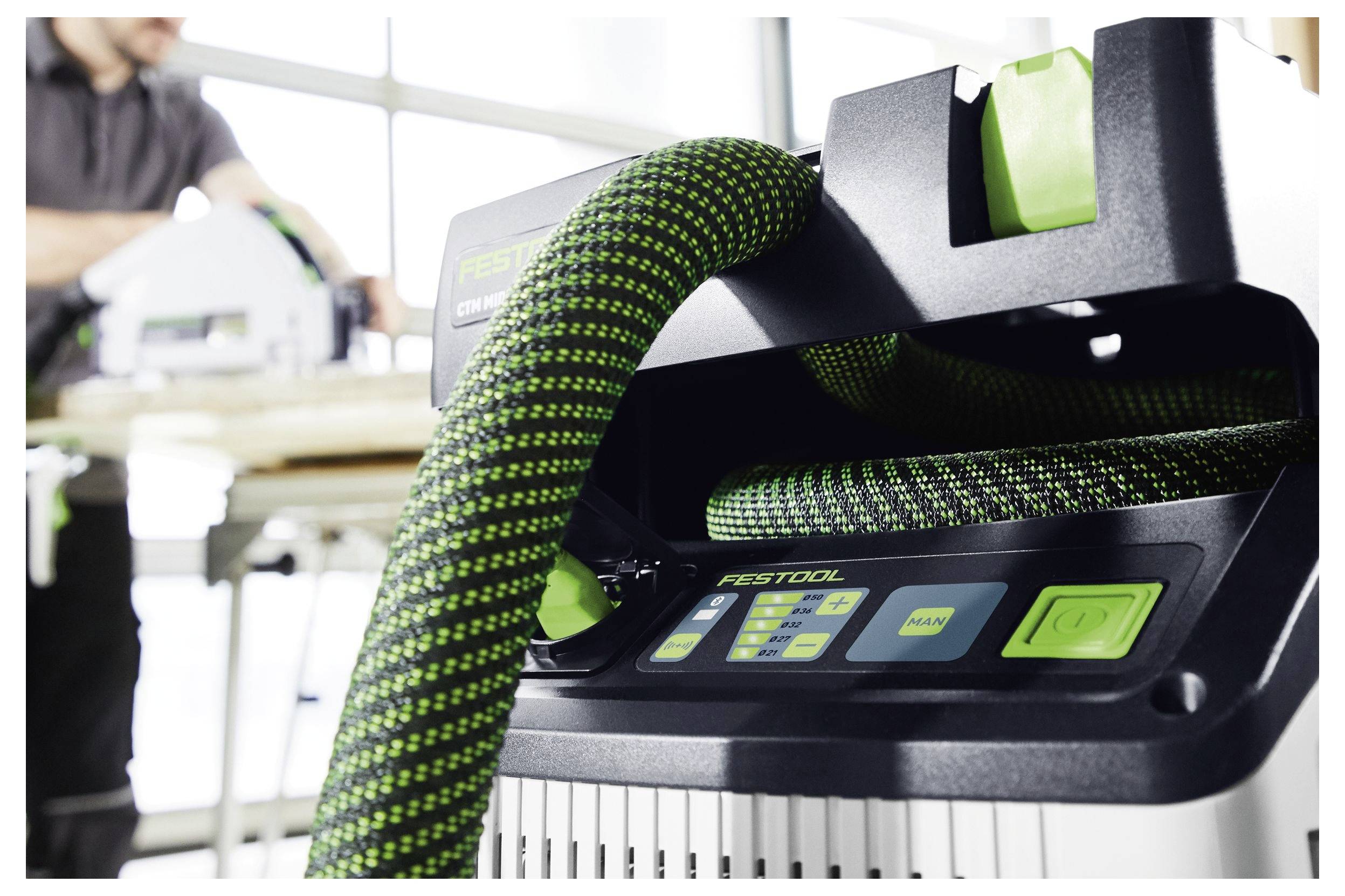 Ein Staubsauger mit einem grünen und schwarzen Schlauch ist im Fokus, während eine Person im Hintergrund in einer Werkstatt an einer Maschine arbeitet.