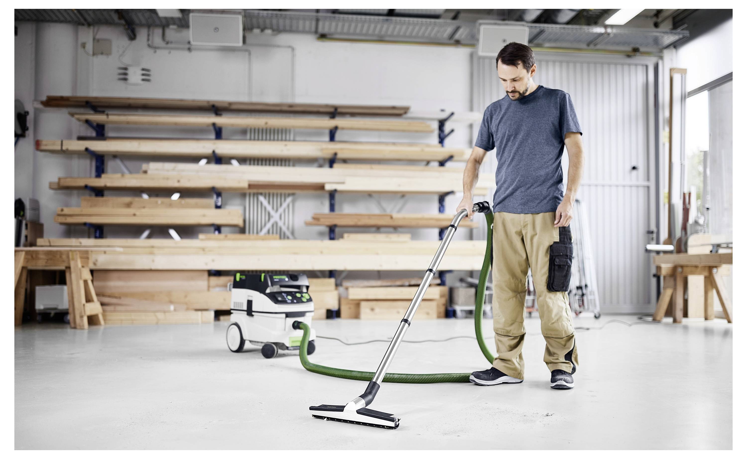 Eine Person, die in einer Werkstatt einen Bodenstaubsauger benutzt, mit Holzbrettern auf Regalen im Hintergrund, wobei Reinigungs- oder Wartungsarbeiten demonstriert werden.