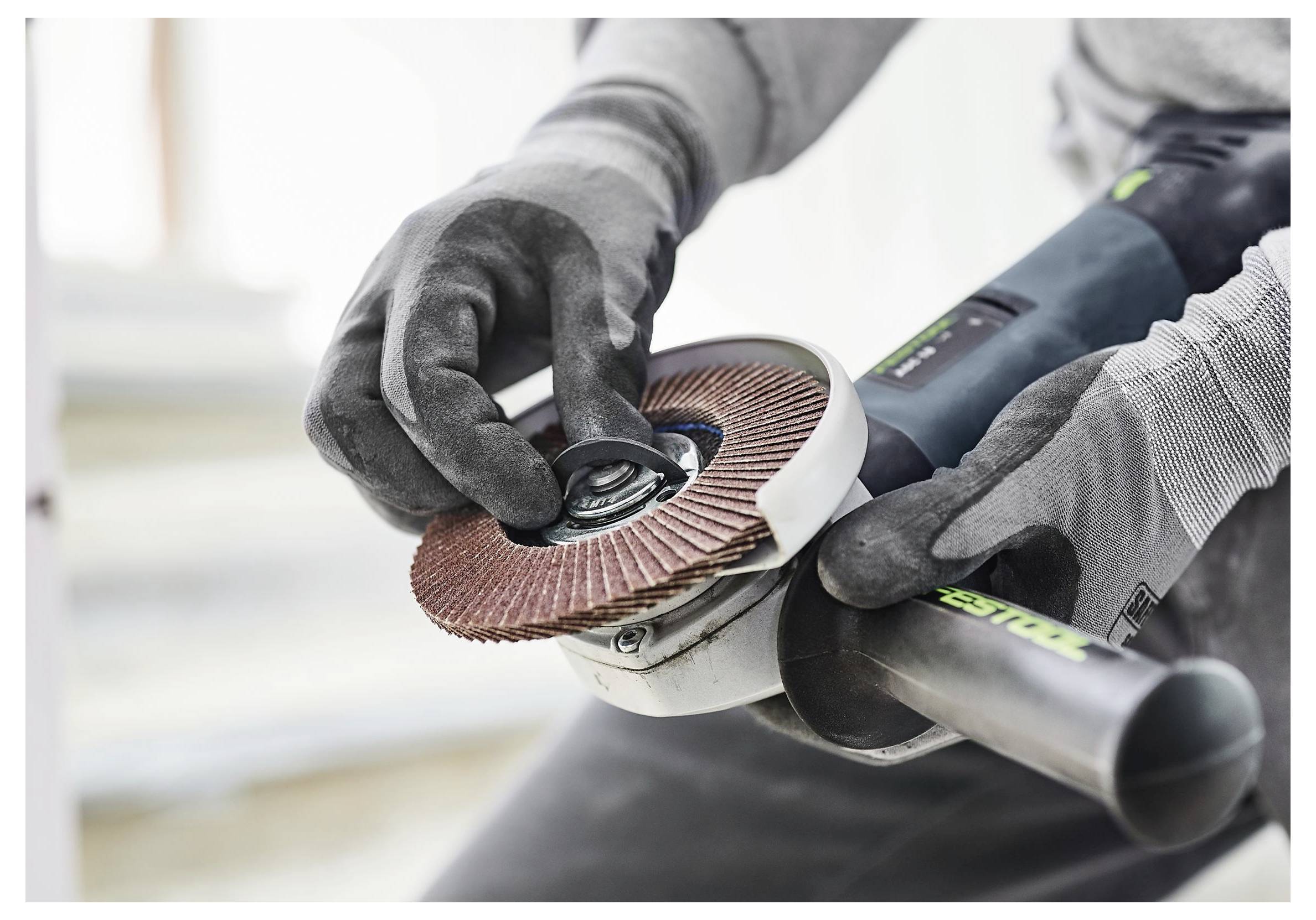 Hände mit Handschuhen, die eine Fächerschleifscheibe an einem Winkelschleifer einstellen, was auf Vorbereitungs- oder Wartungsarbeiten mit Elektrowerkzeugen hindeutet.