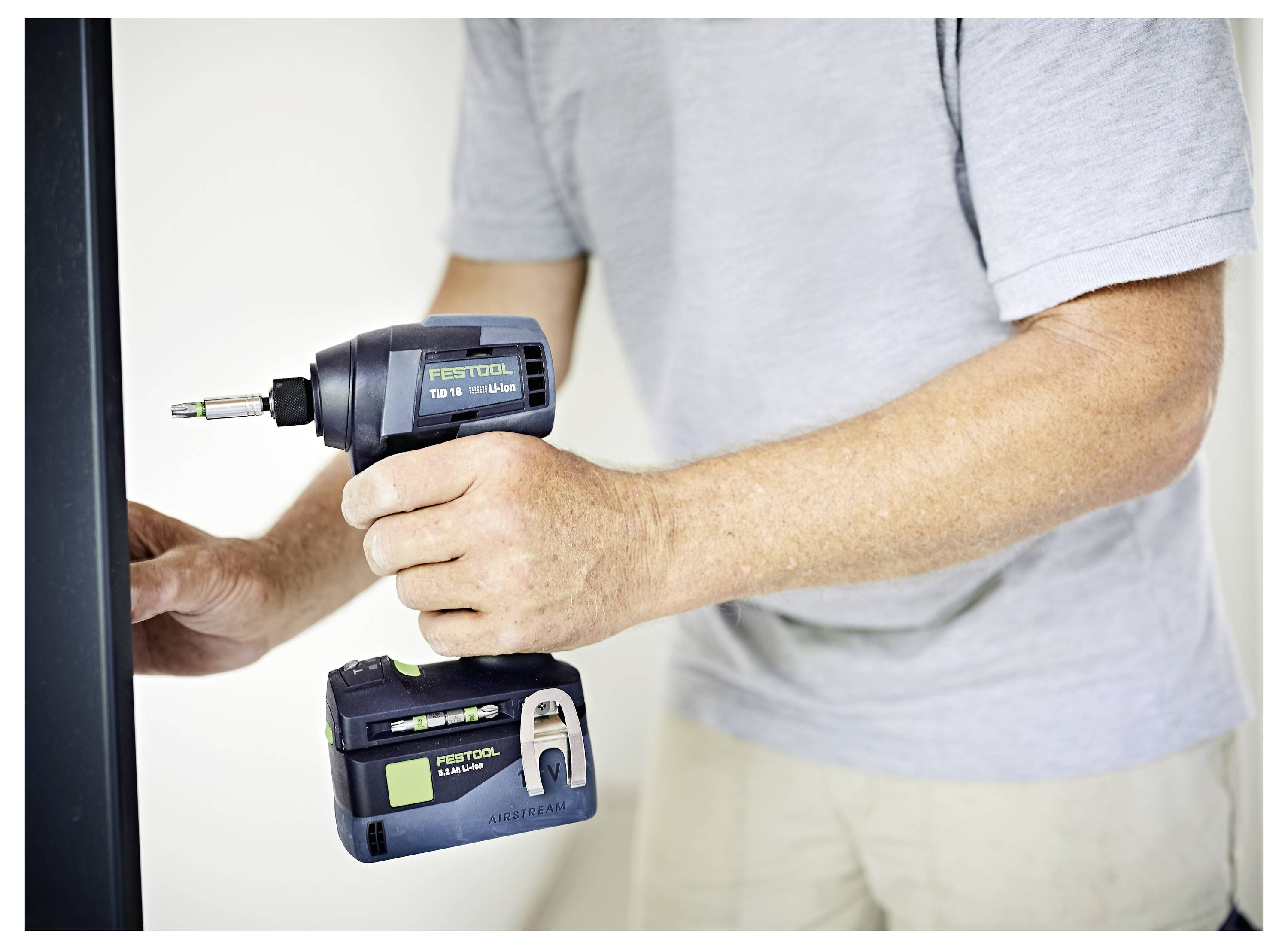 Eine Person, die einen tragbaren elektrischen Bohrer auf einer vertikalen Fläche in einem Raum benutzt. Der Bohrer trägt die Marke 'Festool'.