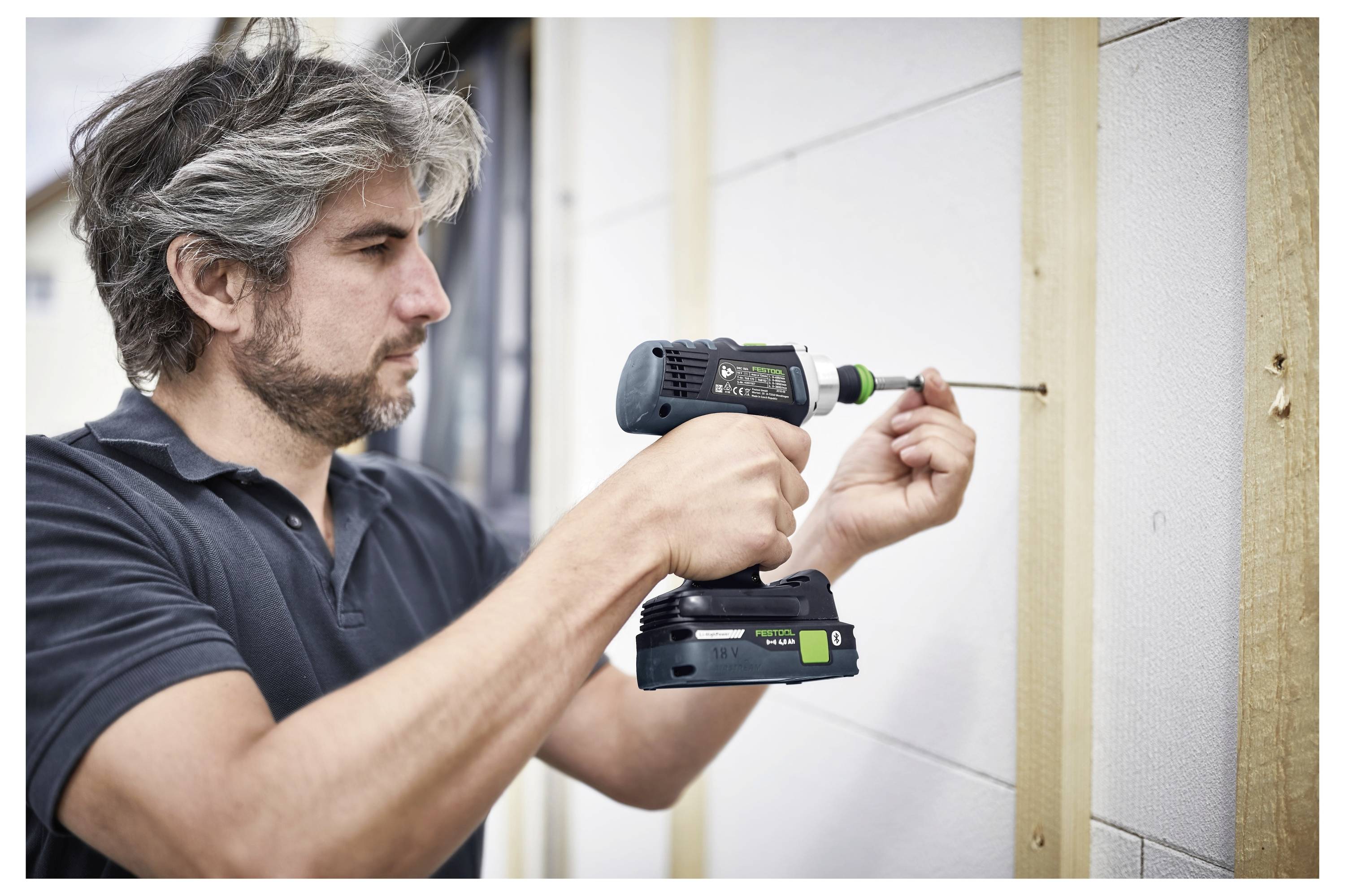 Ein bärtiger Mann benutzt einen kabellosen Bohrer, um eine Holzplatte an eine weiße Wand zu schrauben, wobei er sich konzentriert. Er trägt ein schwarzes Poloshirt.