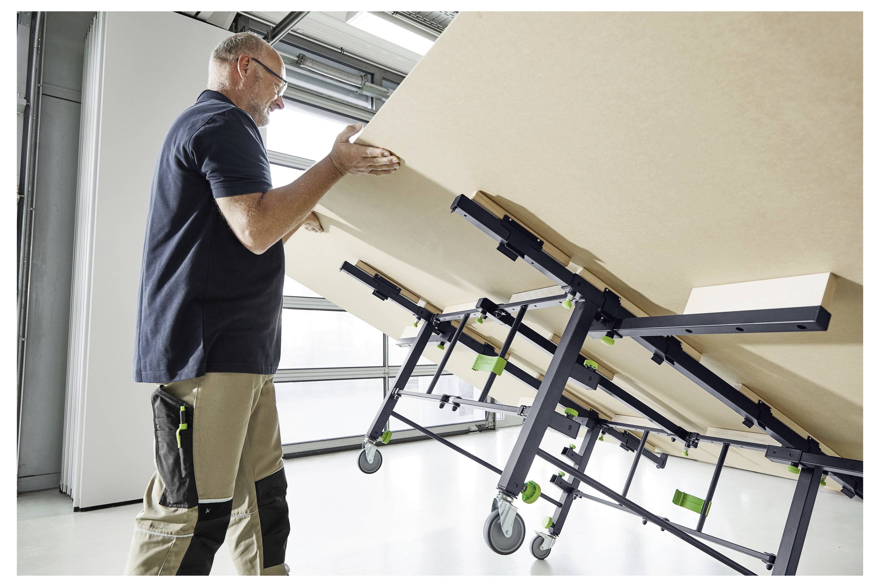 Eine Person hebt ein großes, geneigtes Holzpaneel mit einem mechanischen Gestell mit grünen Rädern in einem hellen Raum mit Glastüren an.