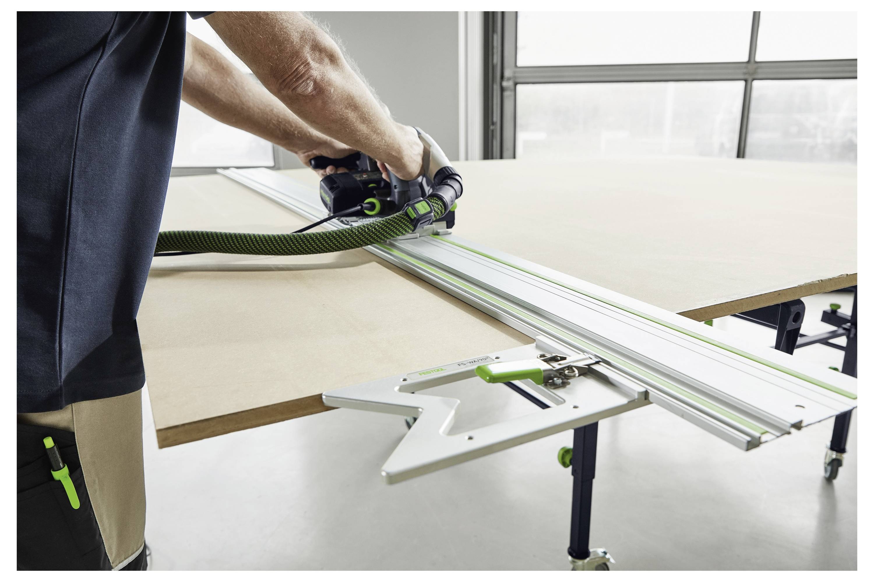 Eine Person benutzt eine Kreissäge mit einer Führungsschiene, um ein großes Holzpaneel auf einer Werkbank in einer Werkstattsumgebung zu schneiden.