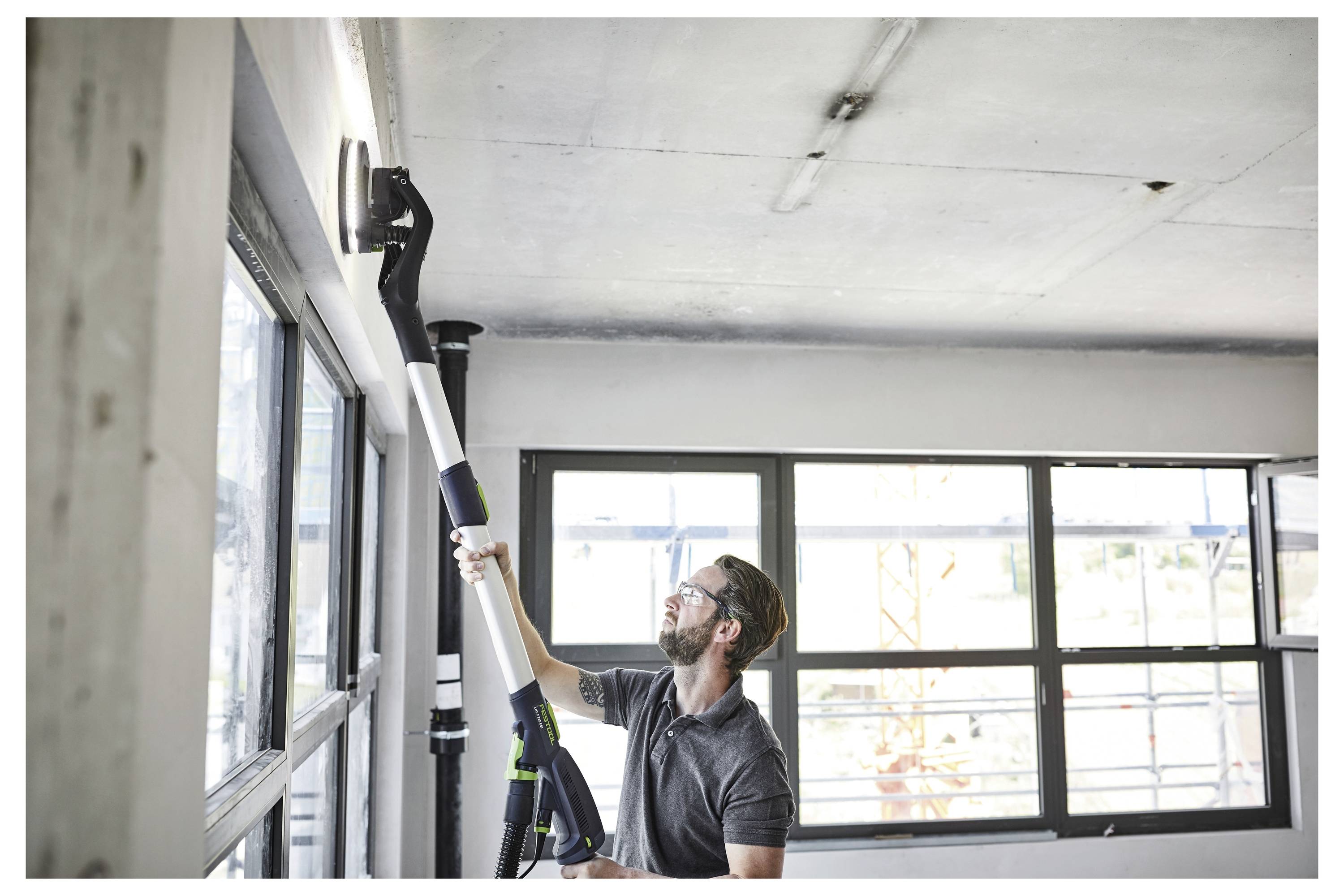 Ein Mann nutzt ein langstieliges Elektrowerkzeug, um die Decke in einem hellen Raum mit großen Fenstern zu glätten.