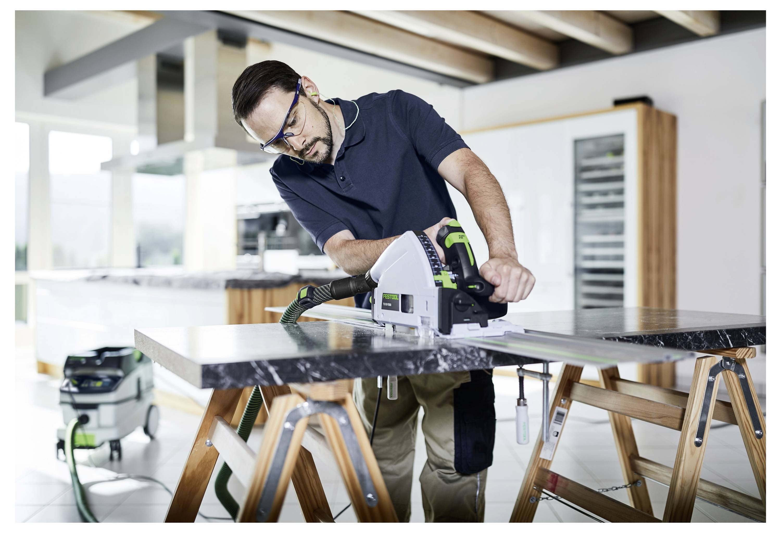 Ein Mann mit Schutzbrille bedient eine Kreissäge auf einer Marmorplatte in einer modernen Küche. Er ist konzentriert und handhabt das Werkzeug sorgfältig.