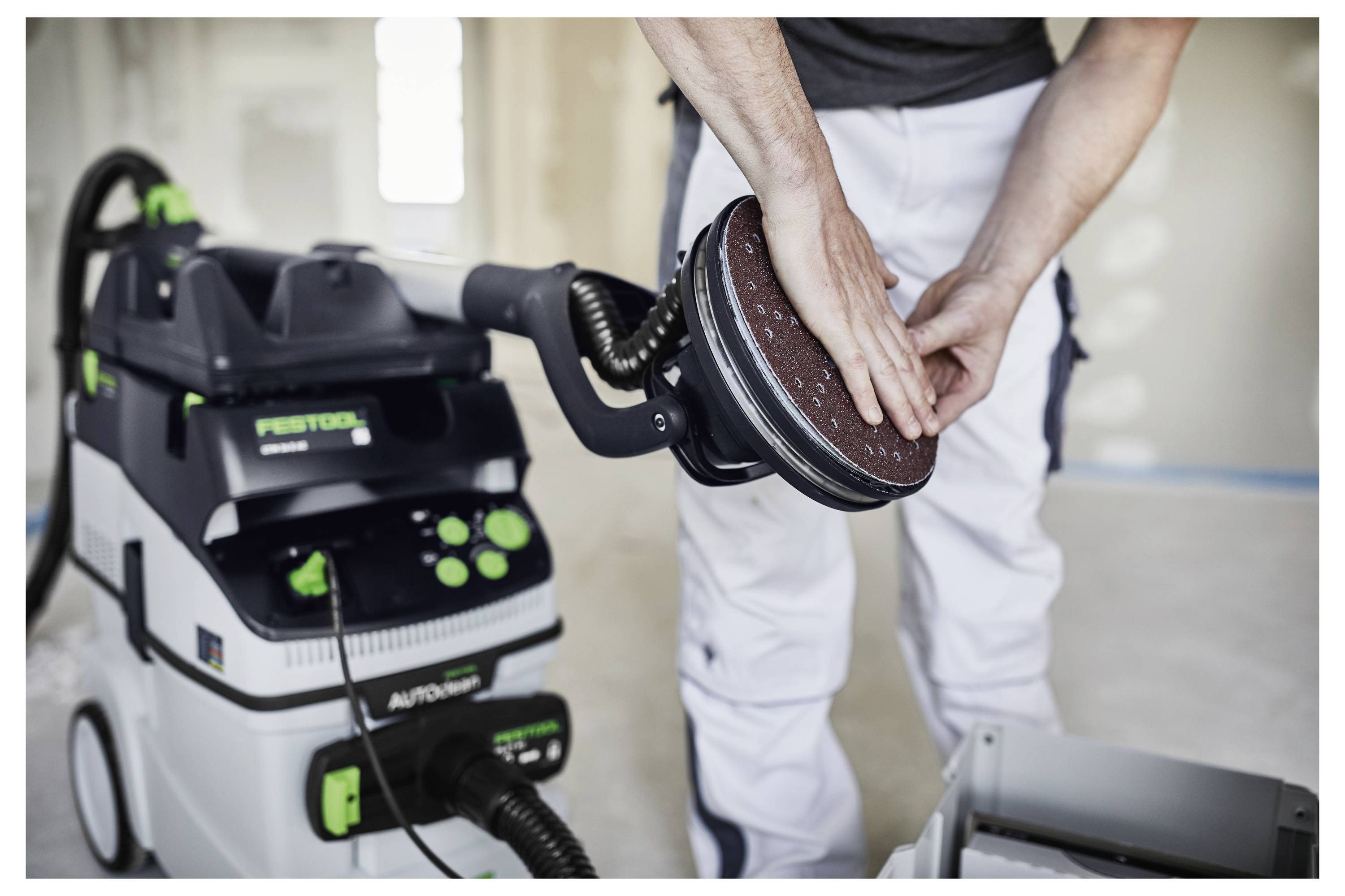 Eine Person in Arbeitskleidung stellt eine Schleifmaschine mit angeschlossenem Staubsauger ein, was auf eine Renovierungs- oder Trockenbaufinishaufgabe hindeutet.