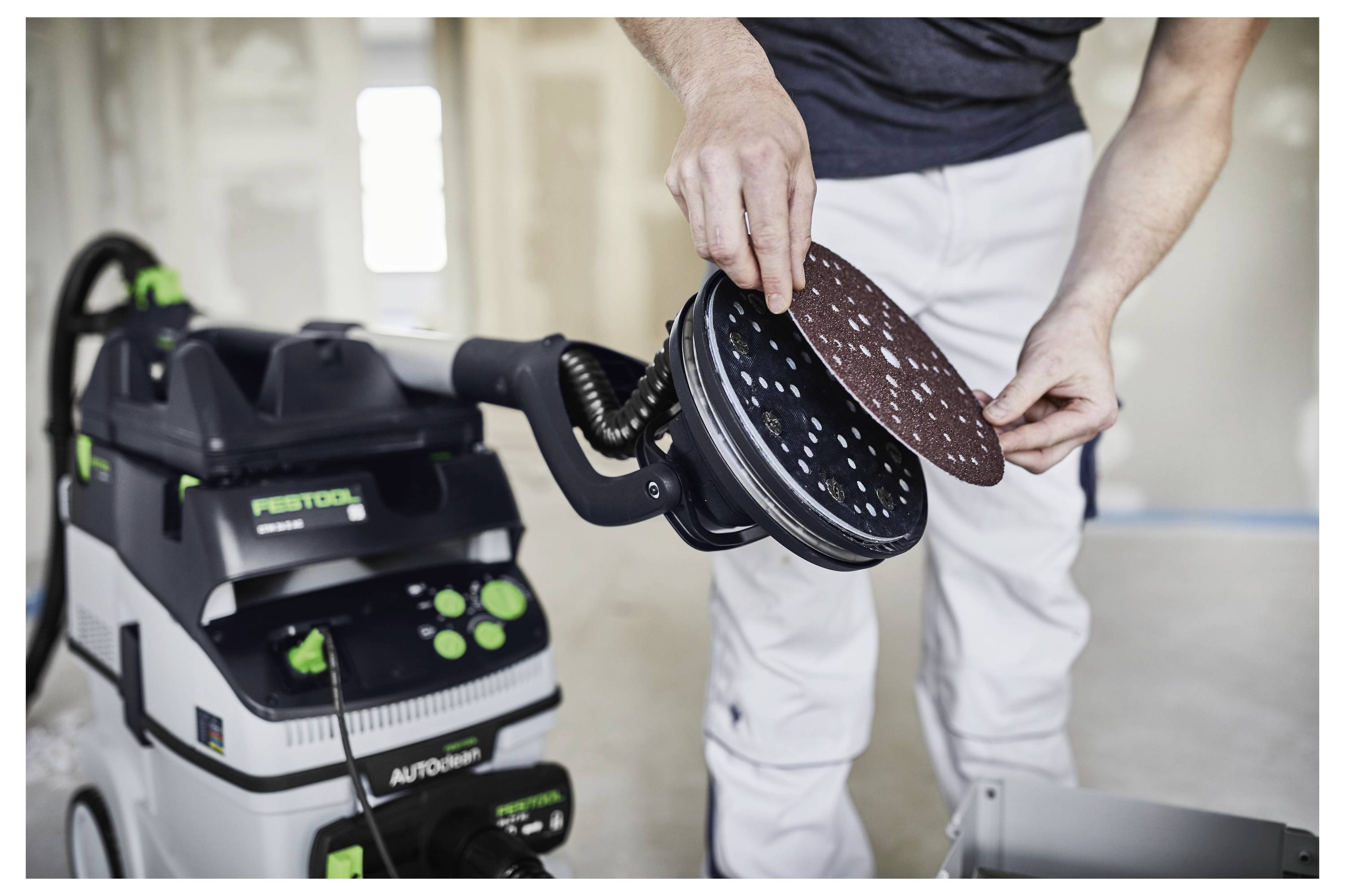 Eine Person befestigt ein Schleifpad an einem Elektrowerkzeug in einer Werkstattsituation. Ein Festool-Staubabsauggerät ist im Hintergrund sichtbar.