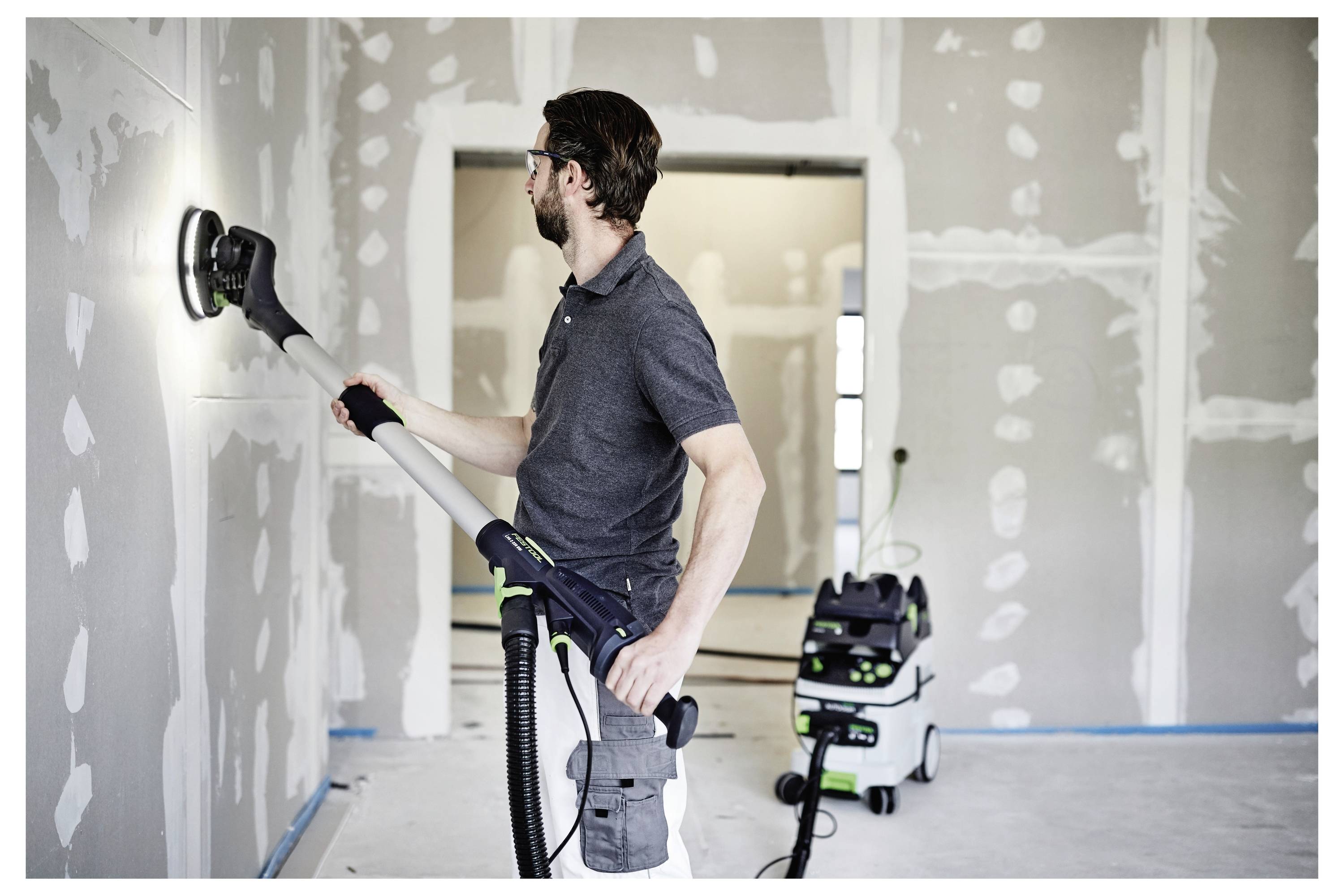 Eine Person schleift Trockenbau mit einem elektischen Schleifers in einem teilweise fertiggestellten Raum. Ein mit dem Schleifers verbundener Staubsauger sammelt Staub.