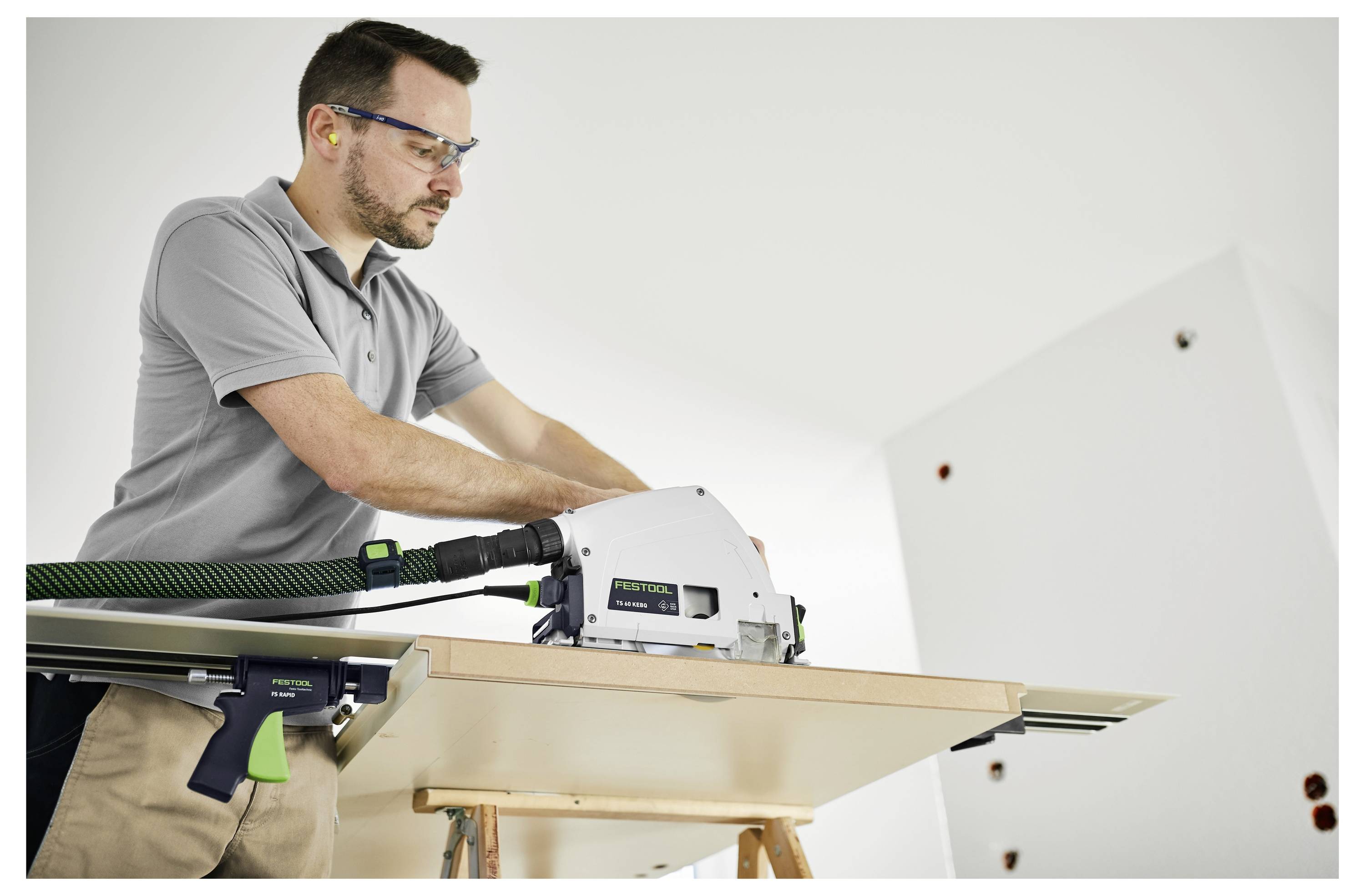 Ein Mann benutzt eine Elektrosäge, um ein Holzbrett zu sägen, während er eine Schutzbrille und einen Gehörschutz in einer Werkstatt trägt.