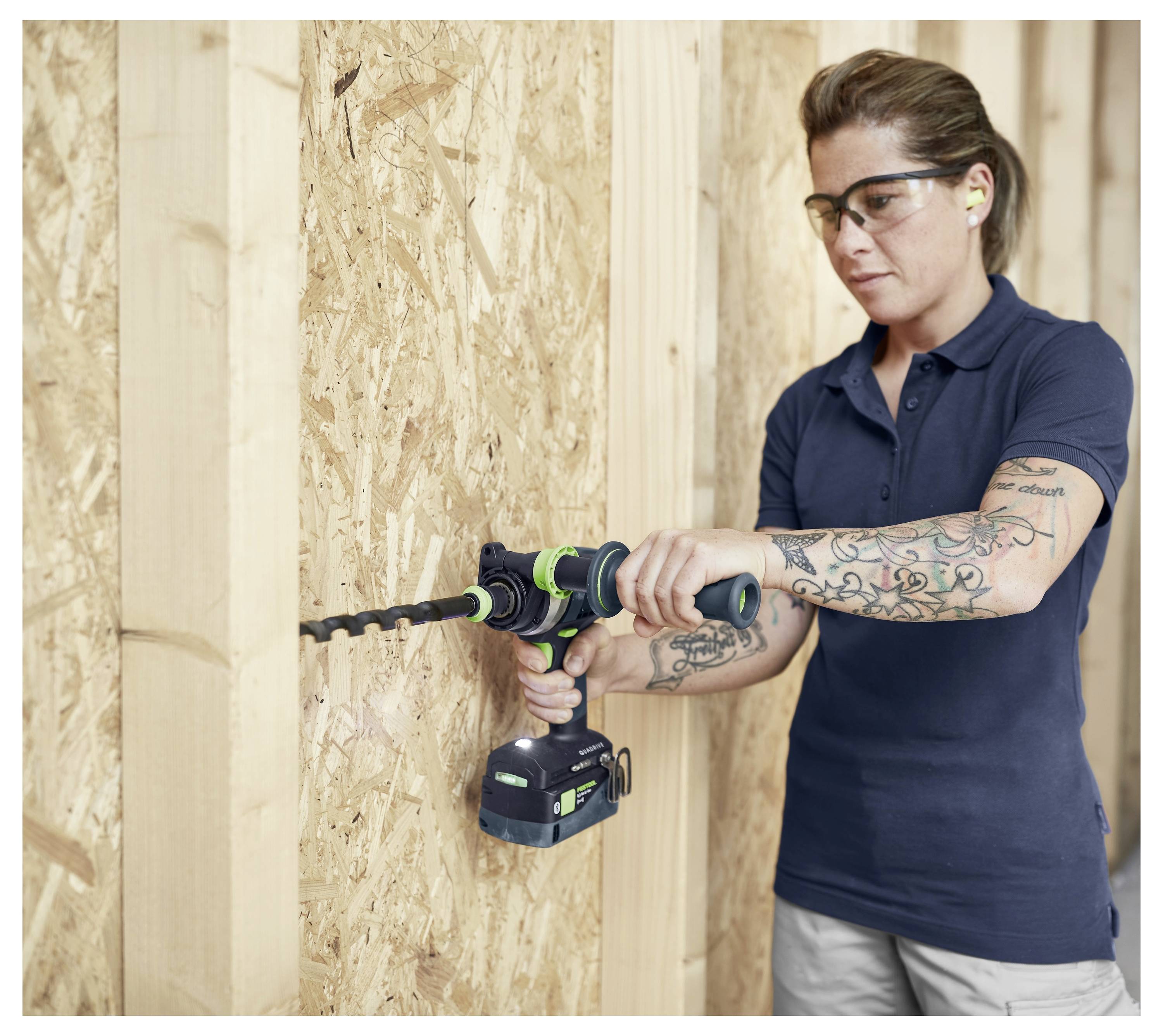 Eine Person trägt Schutzbrille und Gehörschutz und benutzt einen Elektrobohrer an einer Holzwand, wobei sie sich intensiv auf die Aufgabe konzentriert.