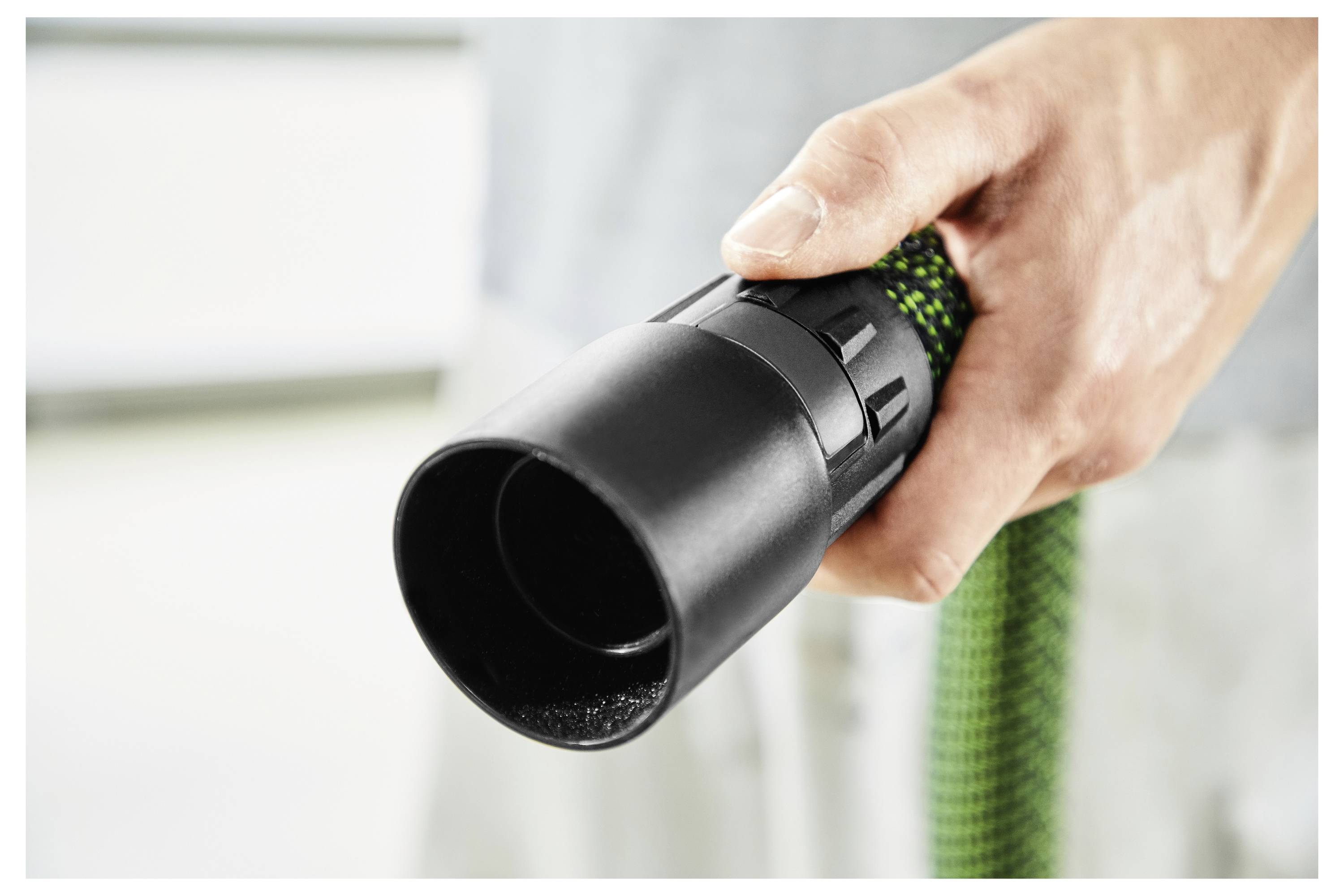 A hand holding the nozzle of a green and black vacuum hose, ready for use, on a blurred background.