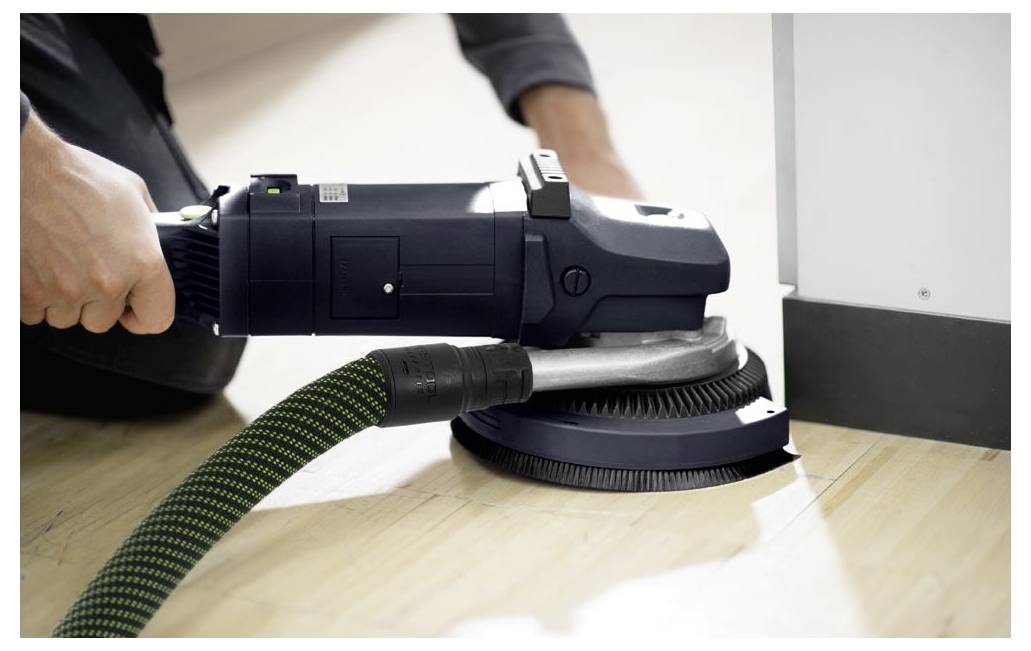 Eine Person verwendet einen Elektroschleiffer auf einem Holzboden neben einer Wand und hält gleichzeitig einen Staubsaugerschlauch zur Staubabsaugung, wobei der Fokus auf dem Kantenschleifen liegt.